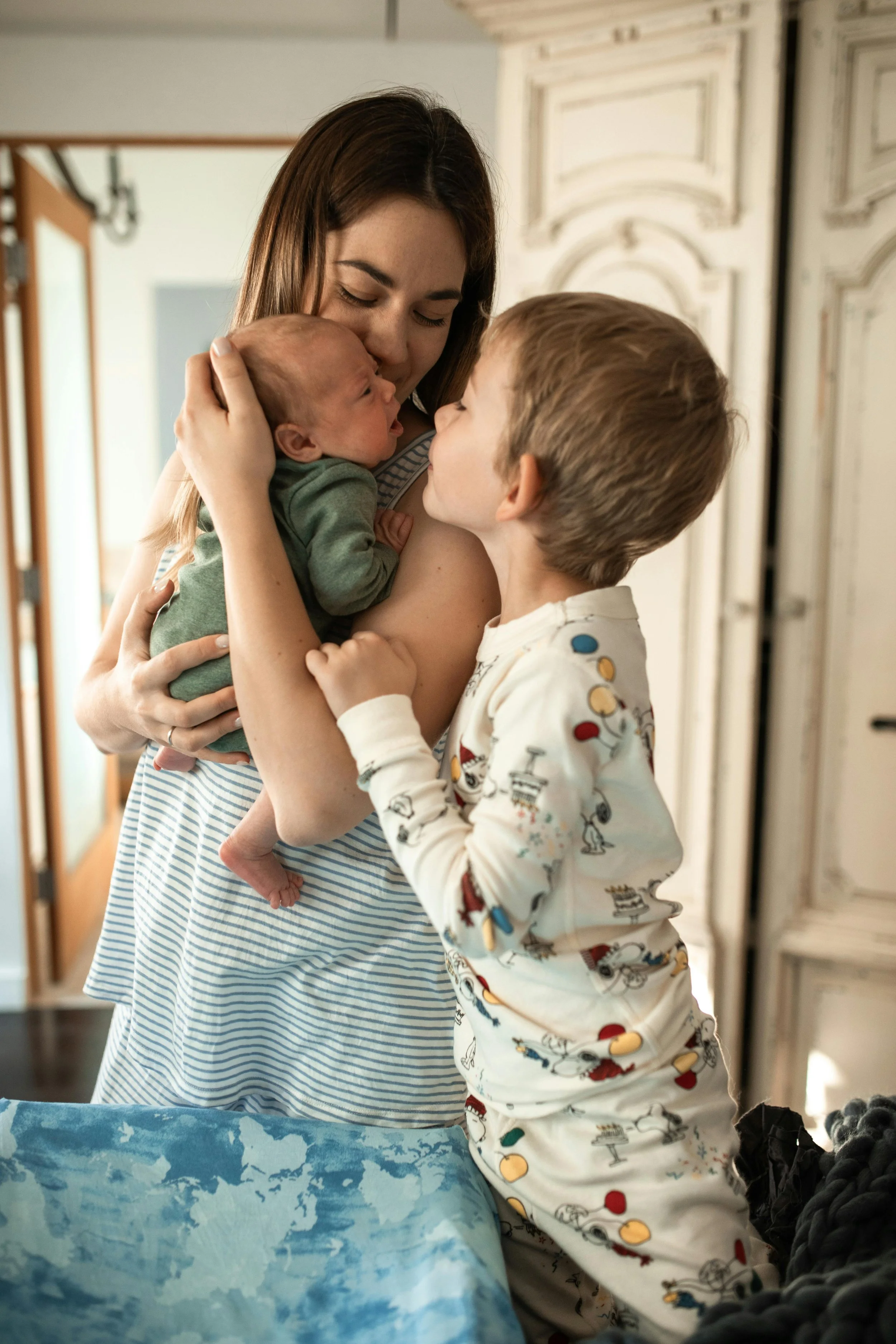 A woman holding a newborn baby in a green outfit while a young boy in pajamas leans in to kiss the baby's forehead in a cozy home setting.