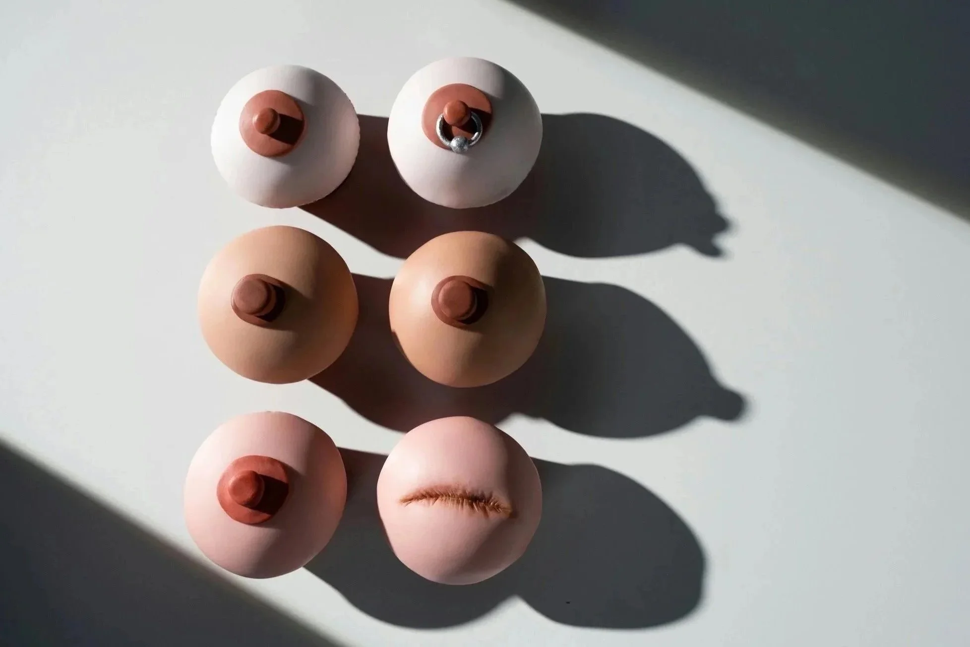 Six breast shaped sculptures of different colors arranged on a white surface with shadows cast to the right.