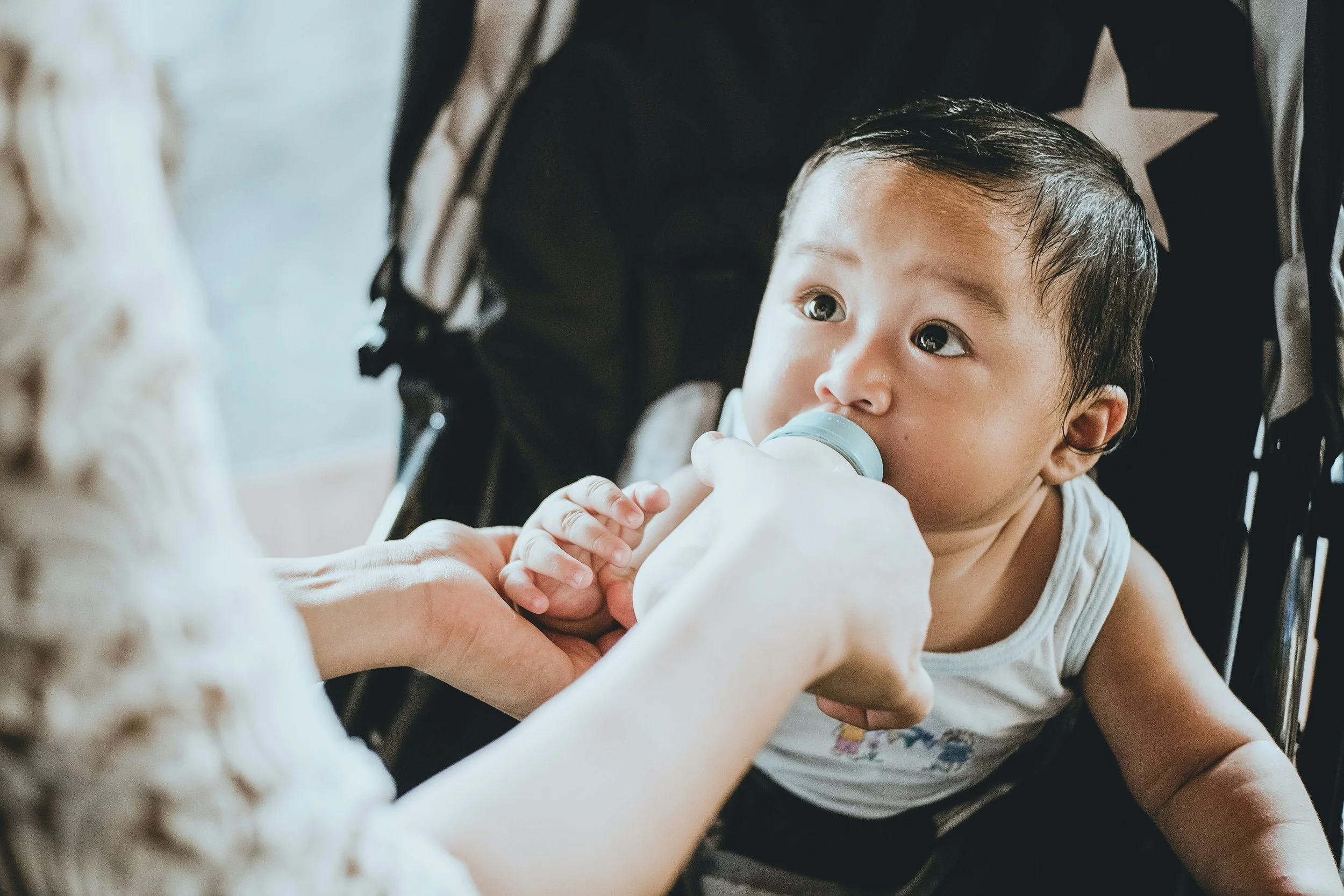 A young child with dark hair sitting in a stroller, being fed with a bottle by an adult.