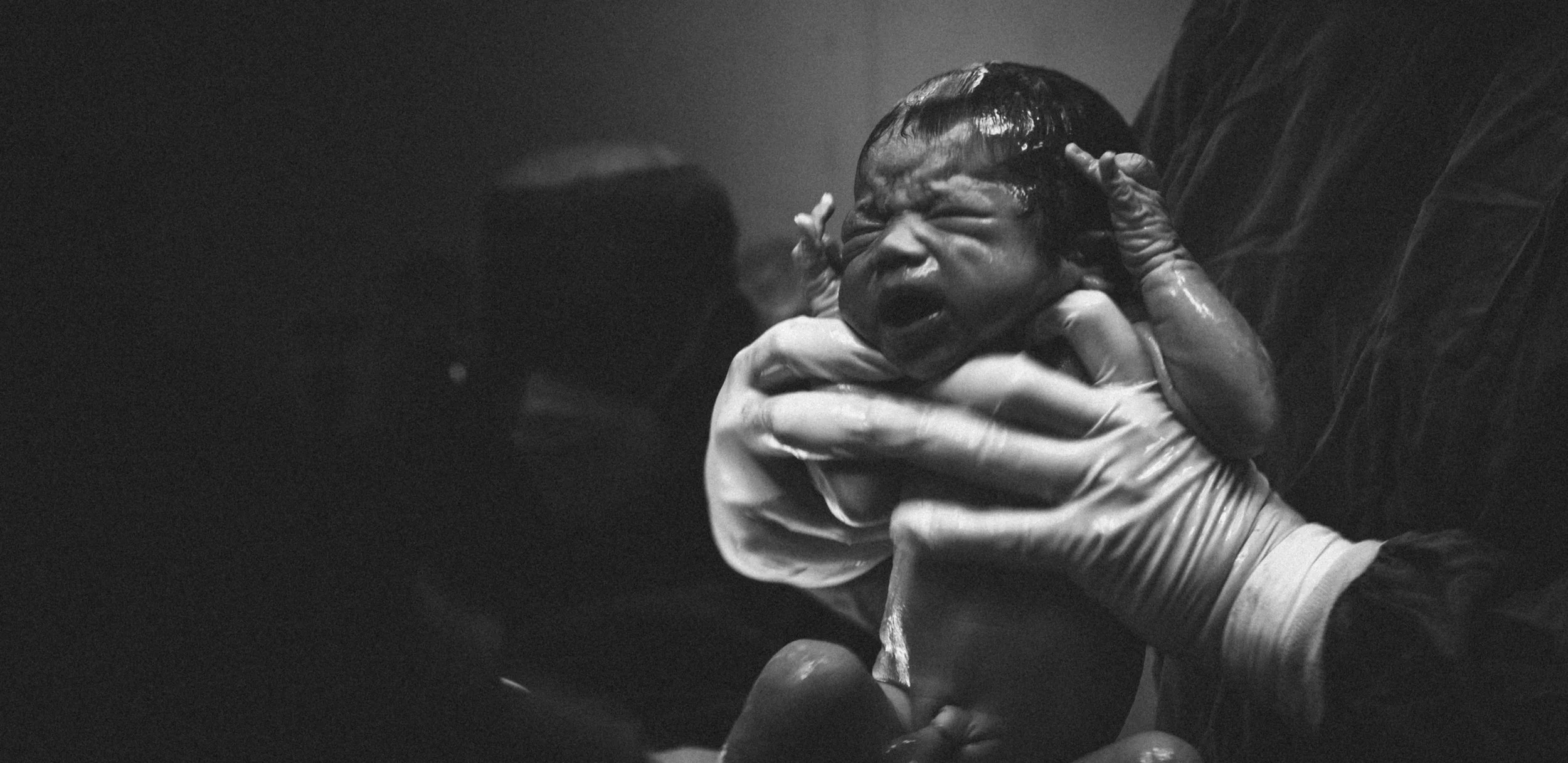 A newborn baby being held by a person wearing gloves, with the baby's face showing an expression of distress or crying.