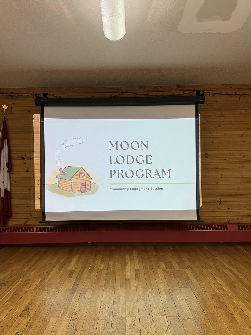 Projection screen displaying a presentation titled "Moon Lodge Program" with a subtitle "Community Engagement Session" and a cartoon illustration of a log cabin emitting smoke.