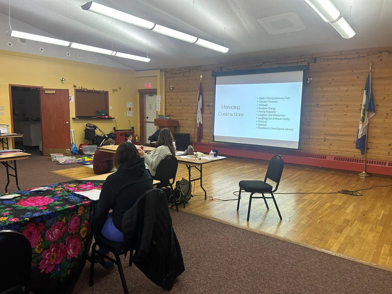 A classroom or seminar room with a projector screen displaying a presentation on managing contractions. Several students are seated around a table, facing the screen. The room has wooden paneling on one wall, yellow-painted walls, and fluorescent lig