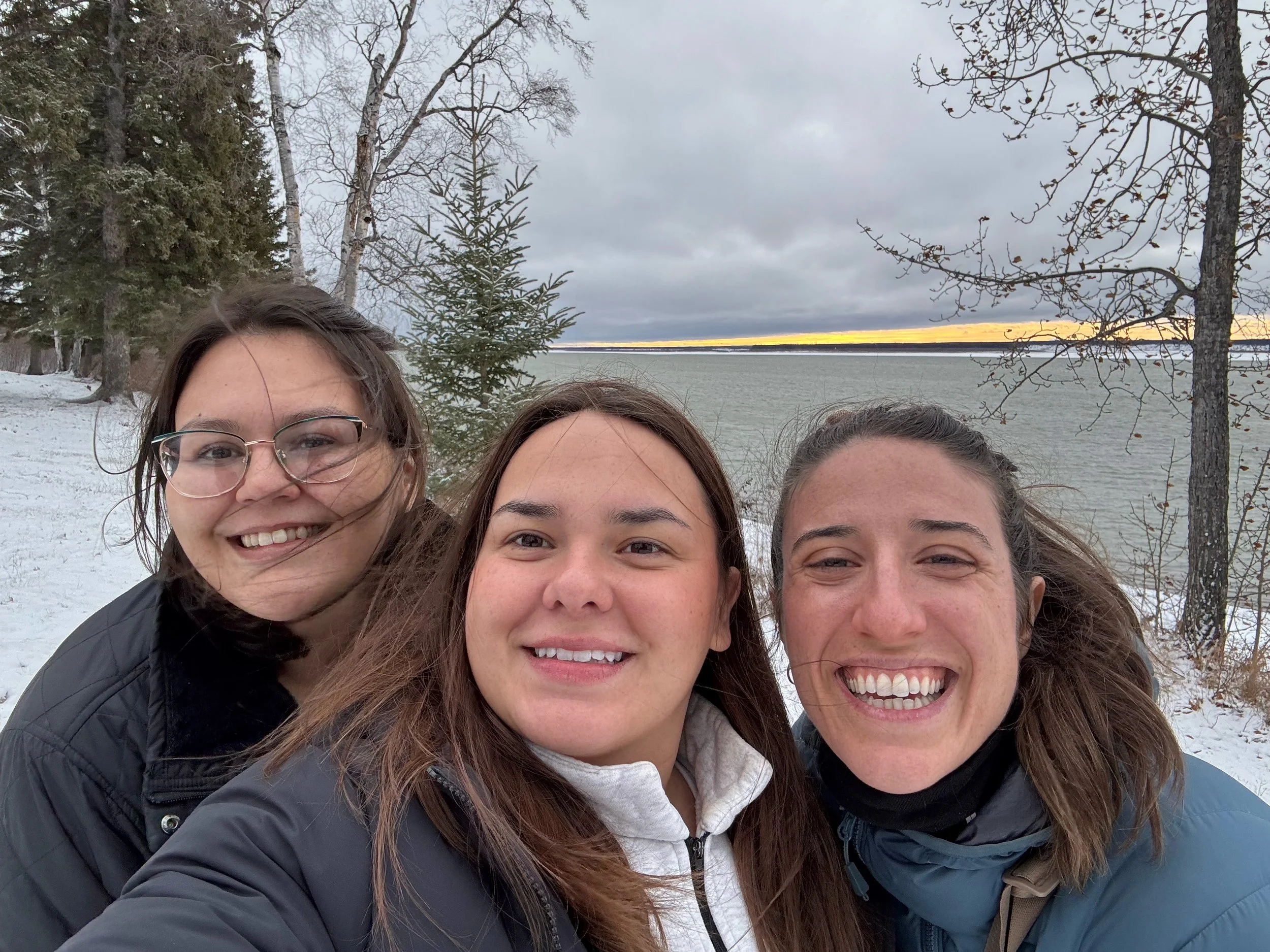 Three women smiling for a selfie outdoors near a body of water with snow-covered ground, trees, and a cloudy sky with a hint of sunset on the horizon.