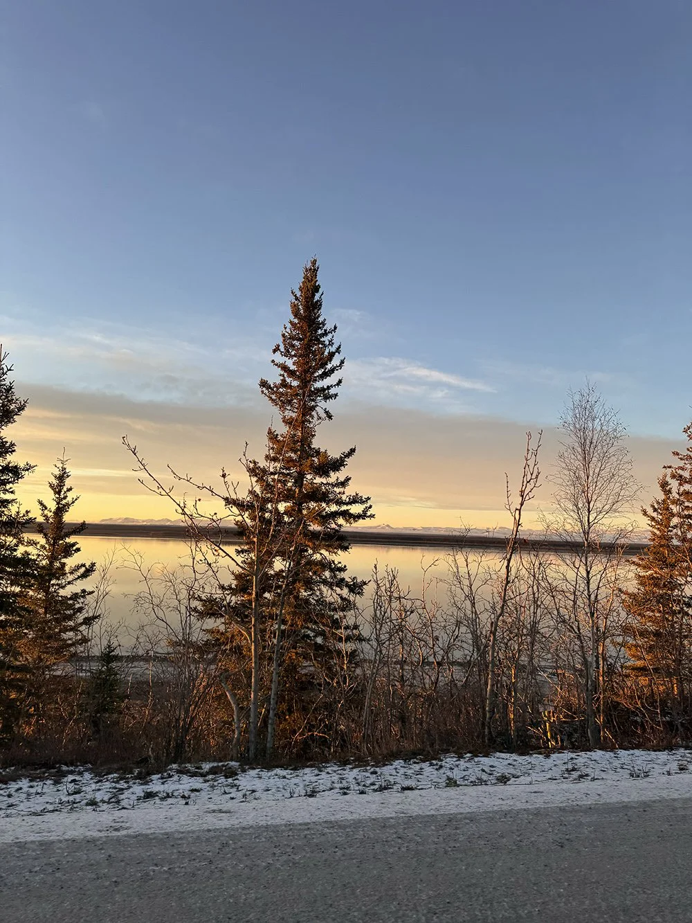 A landscape scene at sunset with a calm body of water reflecting the sky, surrounded by trees, some in early spring without leaves and others evergreen, with snow on the ground.