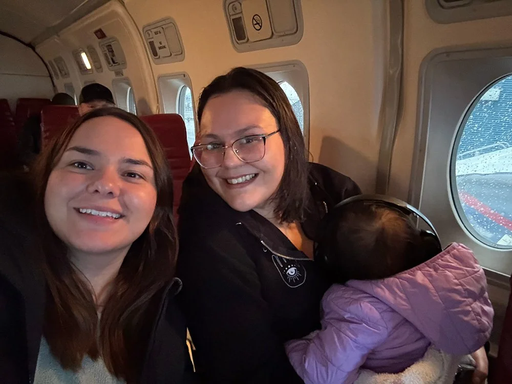 Three women sitting on an airplane, smiling at the camera, with a young child in a pink coat resting on one woman's lap.