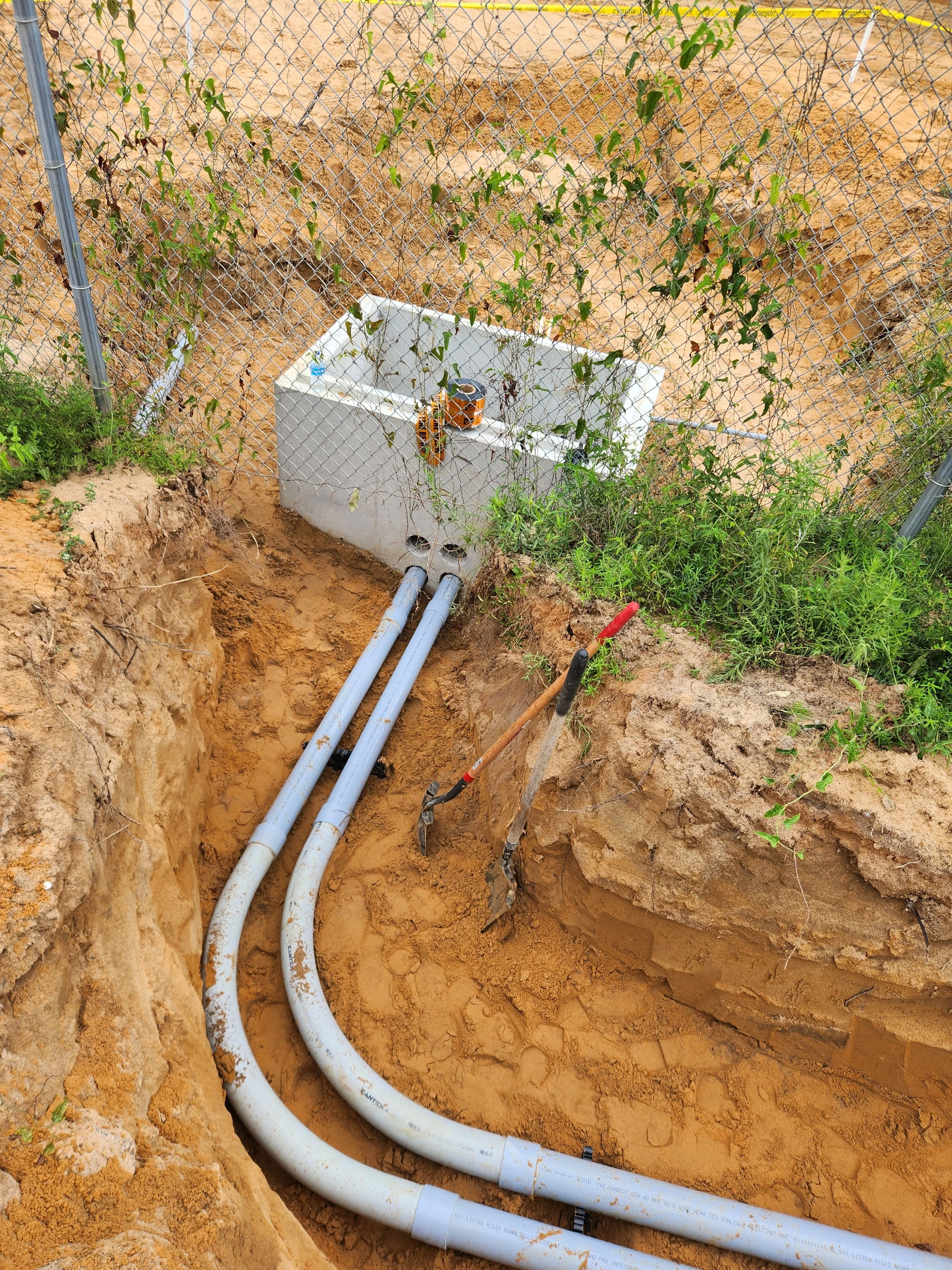 Underground utility pipes and a fitting box in a trench beside a chain-link fence with plants.