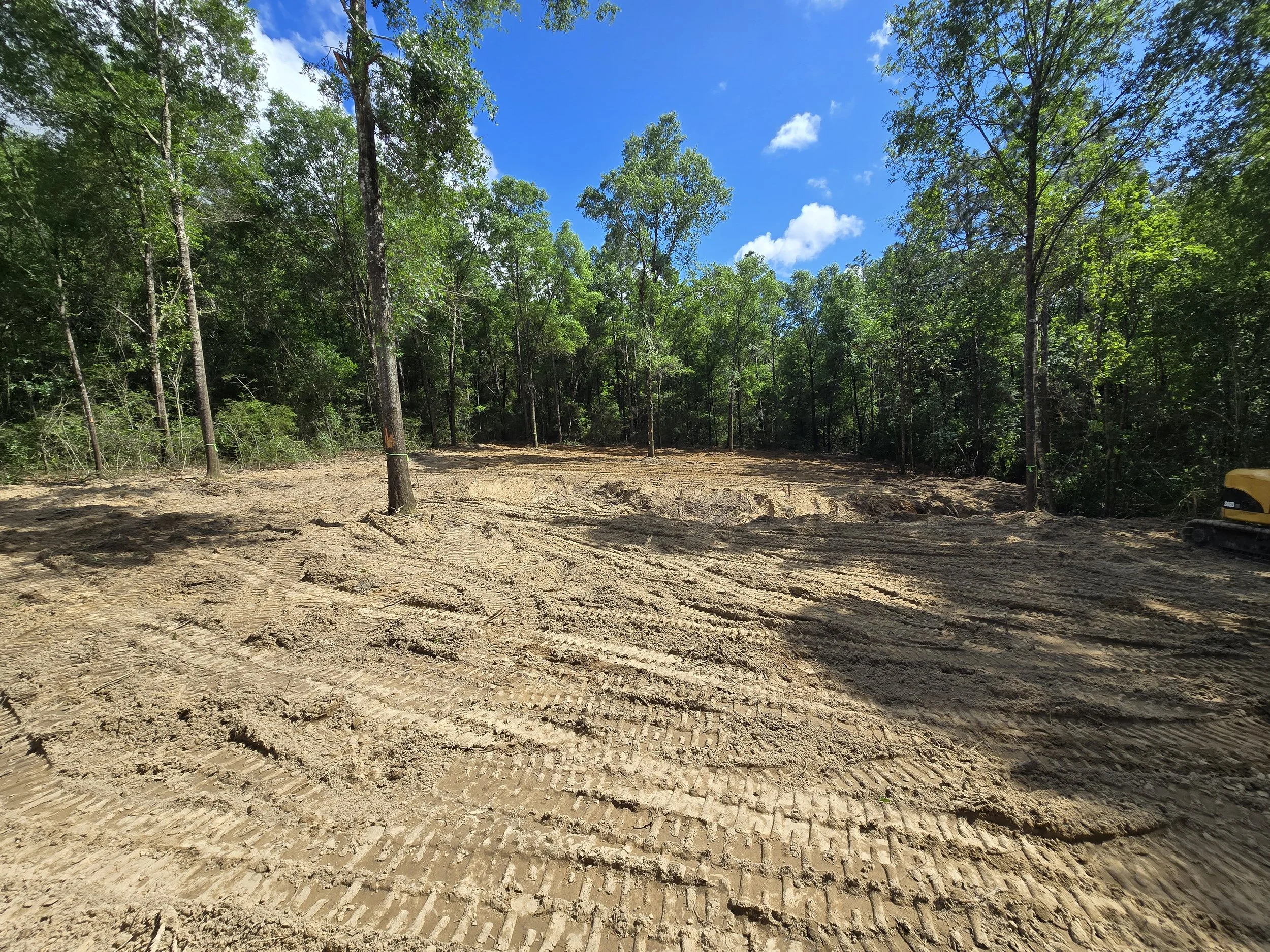 Cleared land with tire tracks, surrounded by trees under a blue sky with scattered clouds.