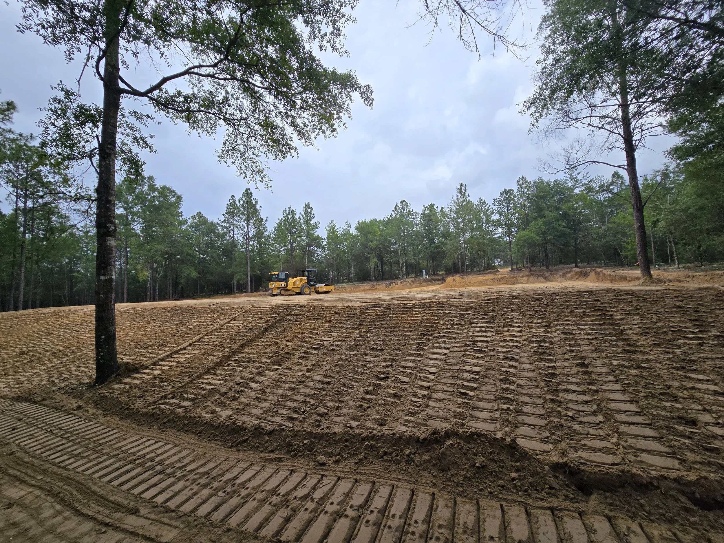 Construction site with freshly leveled and marked ground, a yellow roller compactor in the distance, surrounded by trees and a cloudy sky.