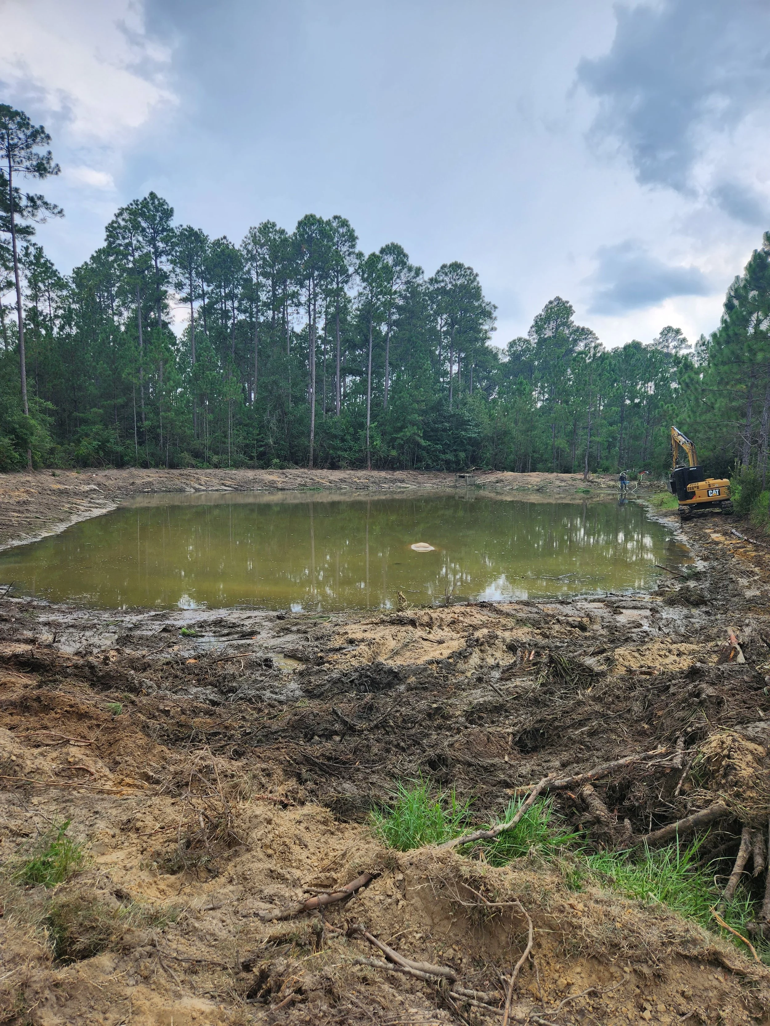 Construction site with a small pond or excavated area, surrounded by dirt and some green grass, with trees and a cloudy sky in the background, and a small yellow excavator on the right side.