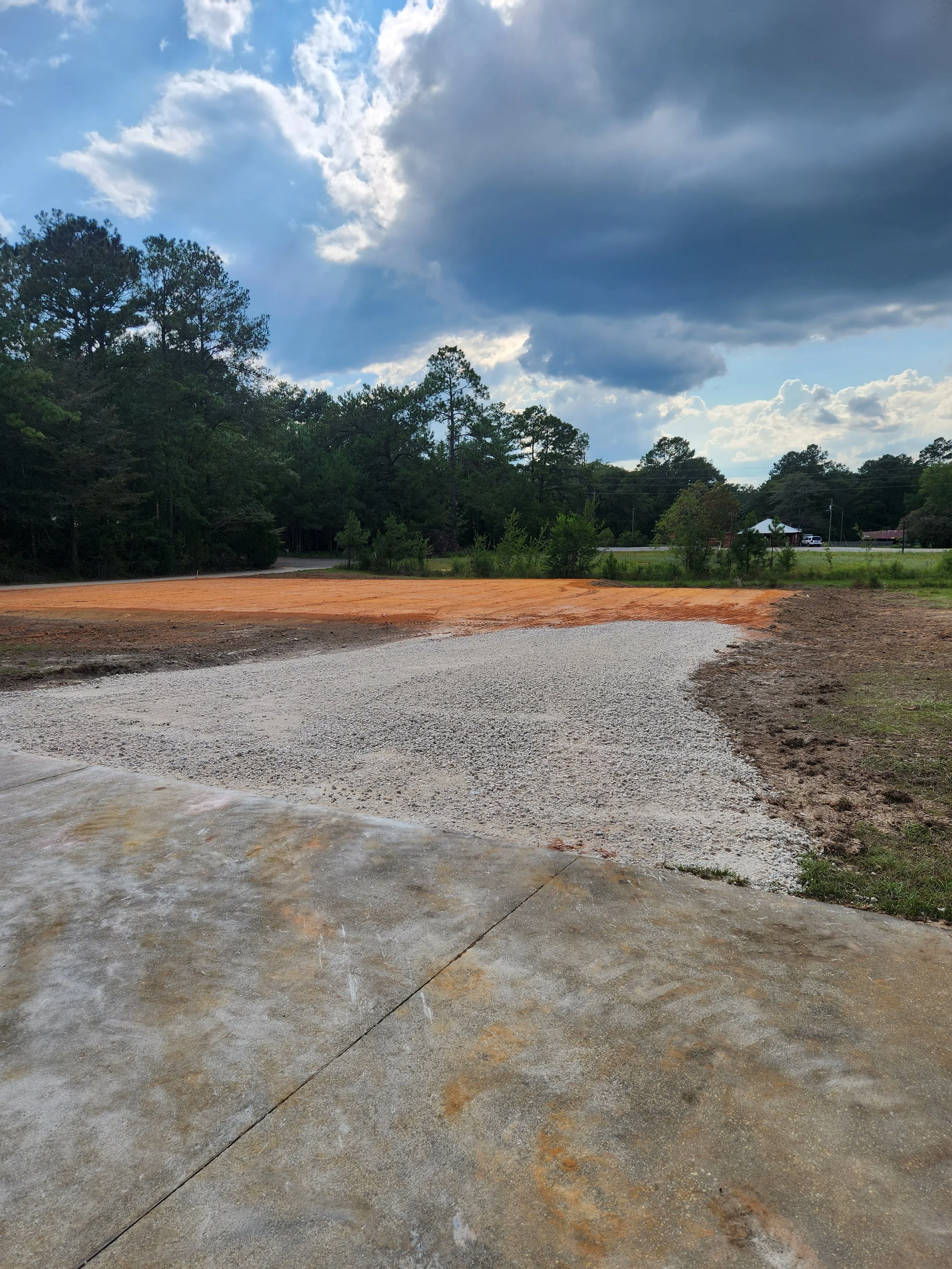 A partly cloudy sky with sunlight peeking through dark clouds over a gravel and dirt parking lot area surrounded by trees and some distant houses.