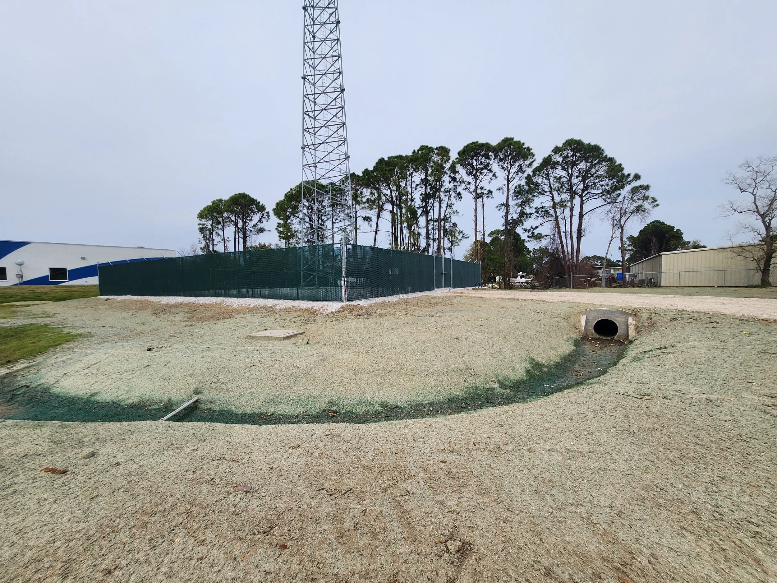 A construction site with a large tower, gravel ground, a drainage pipe, fencing, and trees in the background.