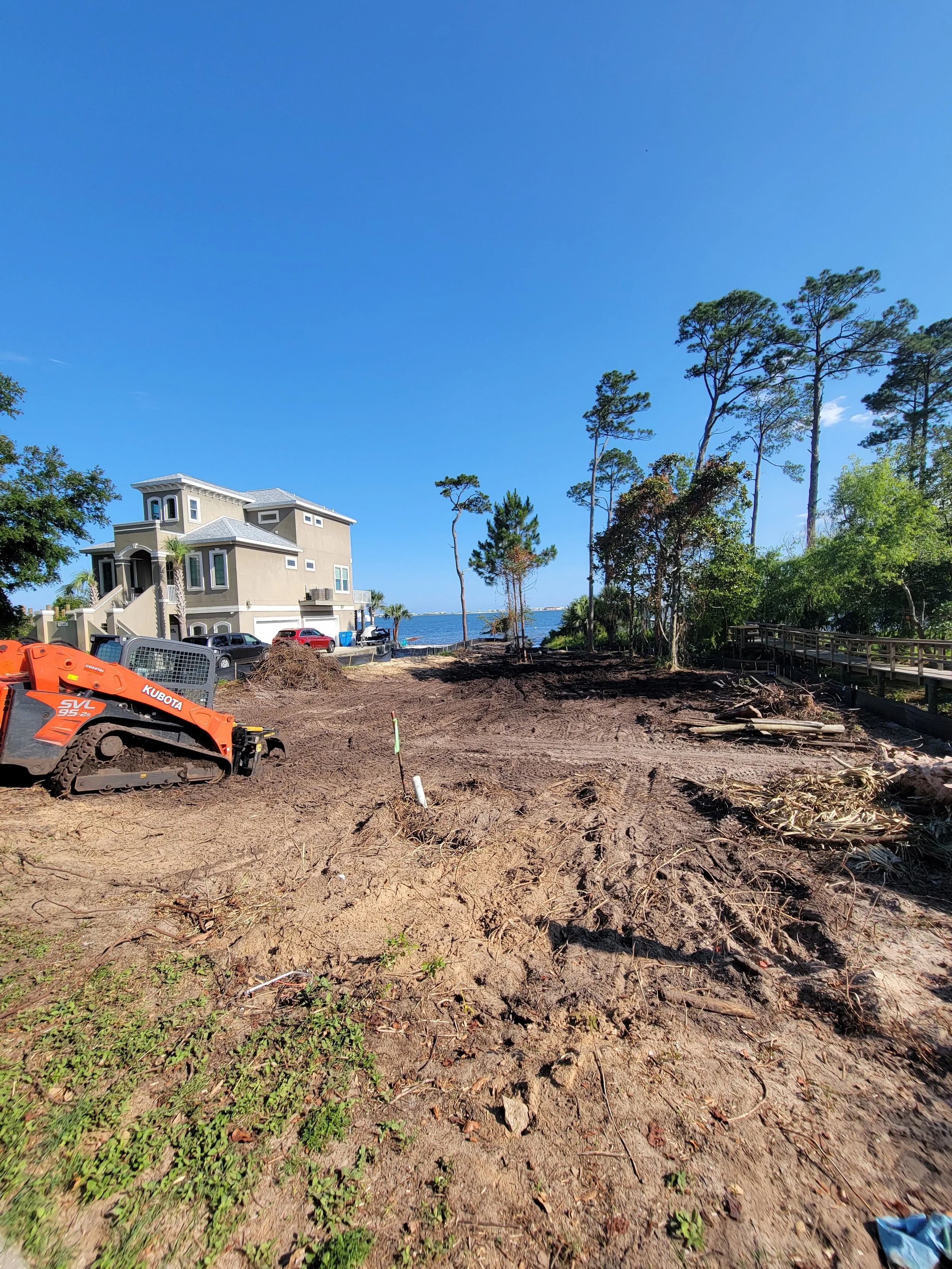 An empty lot with disturbed dirt, a small orange excavator on the left, and a neighborhood with a large house and trees in the background under a blue sky.