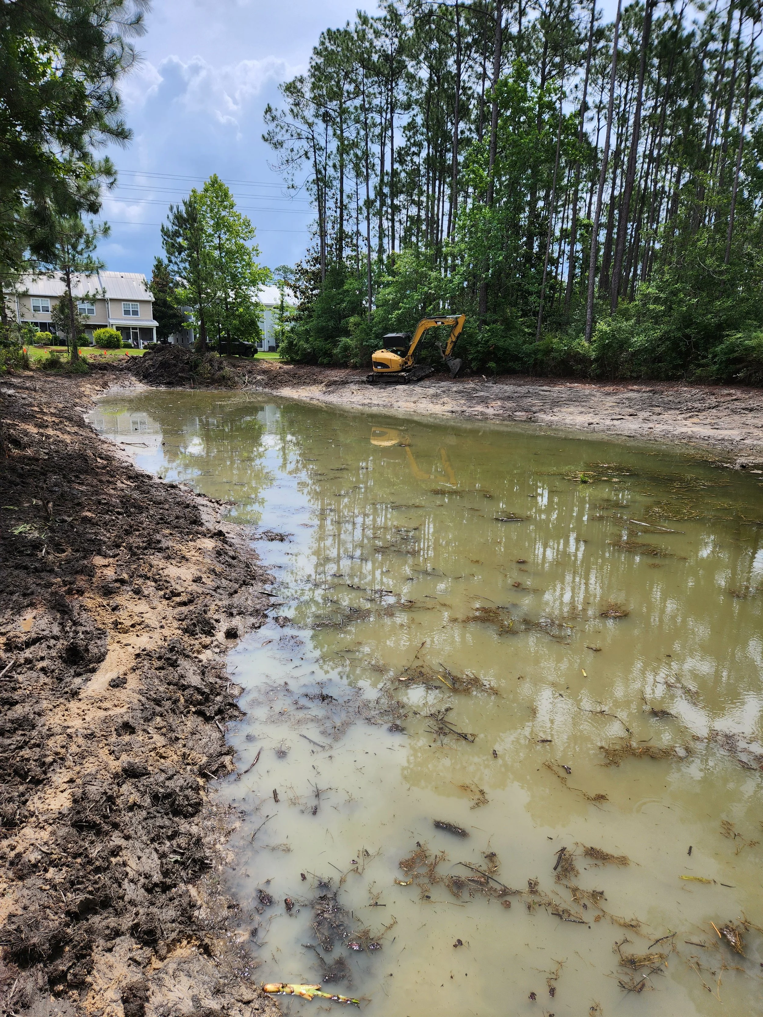 A construction site near a pond with muddy banks and a small excavator, surrounded by trees and residential houses in the background.