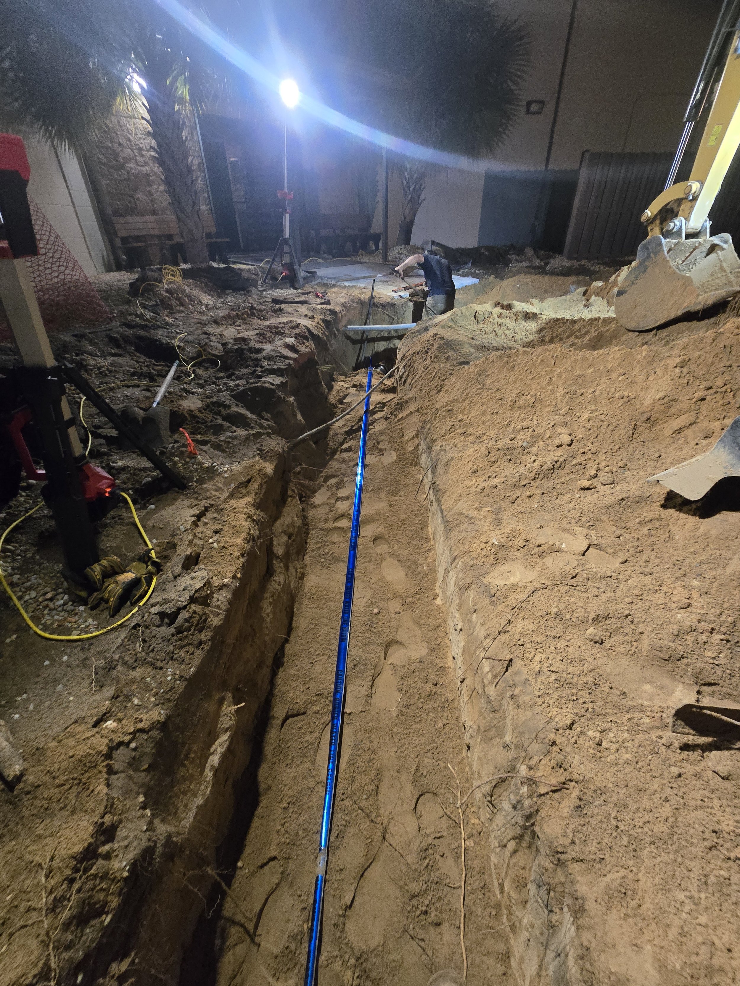 Construction workers are working in an outdoor excavation site at night, with a deep trench, equipment, and a bright light overhead.