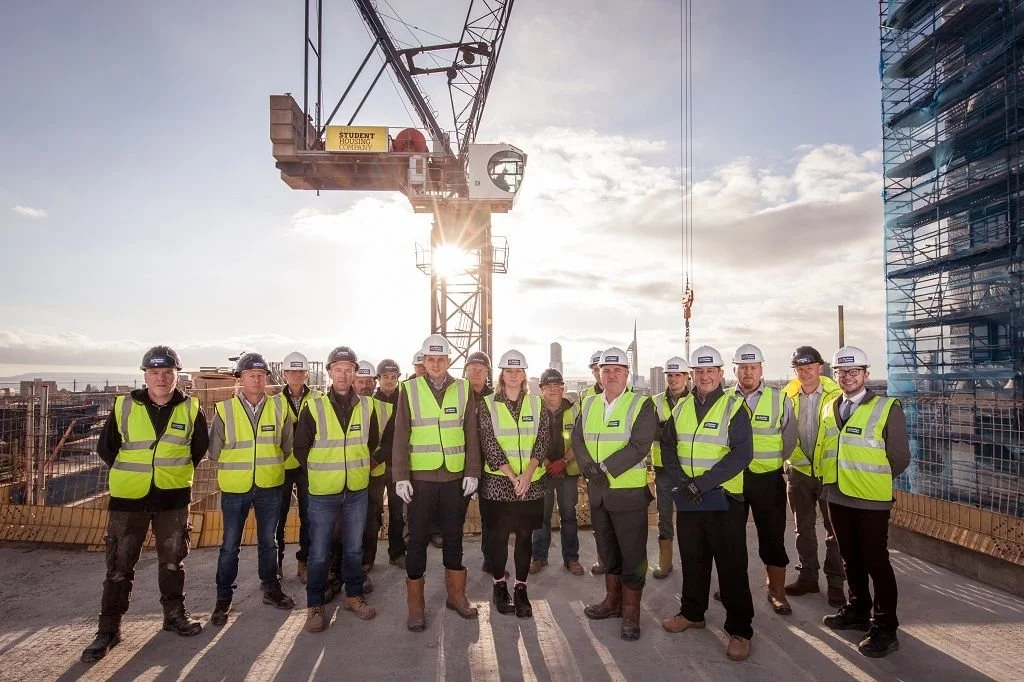 Group of construction workers and professionals in high-visibility vests and helmets on a building site, with a crane and city skyline in the background.