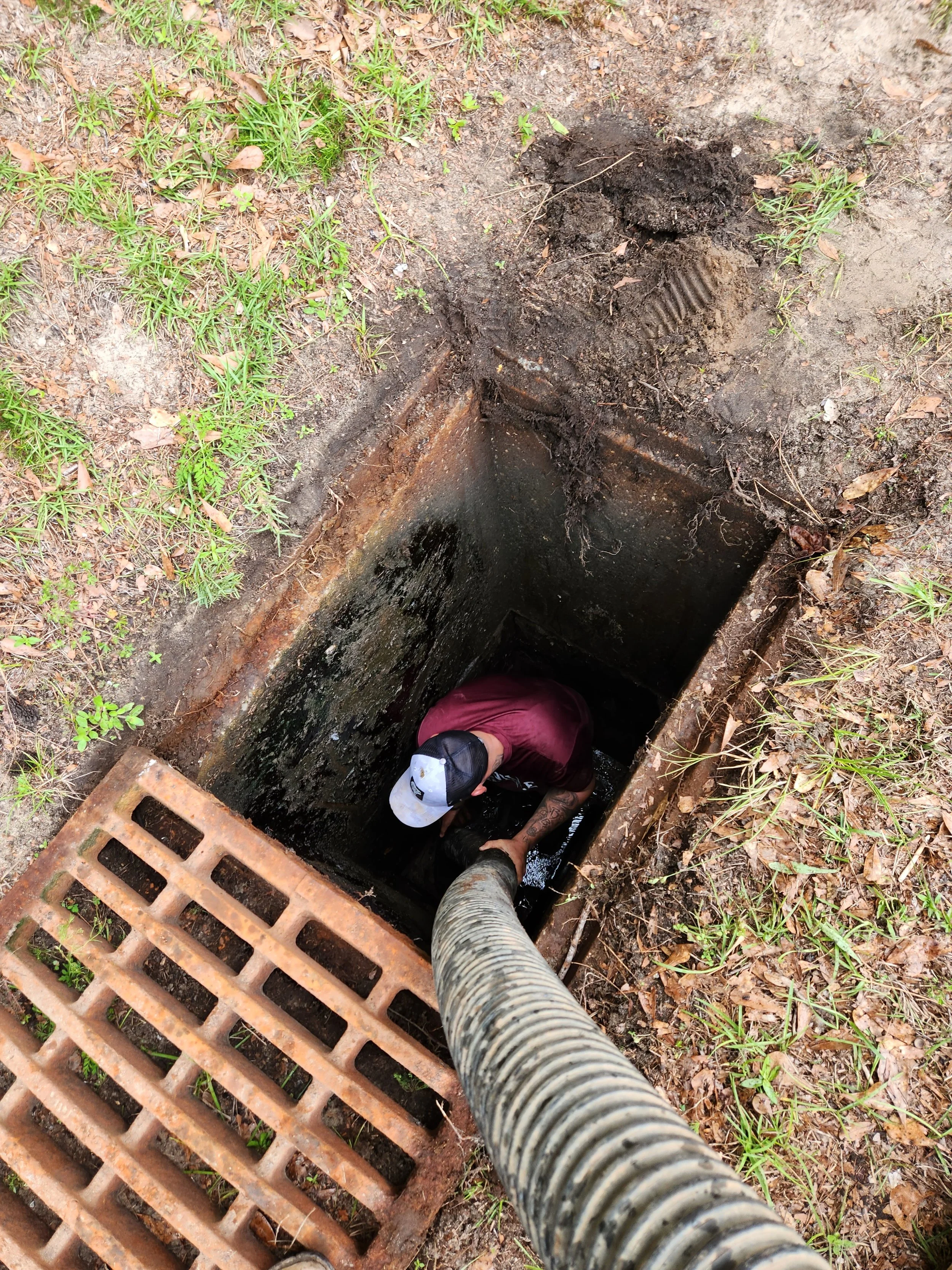 A person wearing a cap and dark clothing is cleaning out a storm drain or drainpipe with a flexible hose, surrounded by grass and dirt.