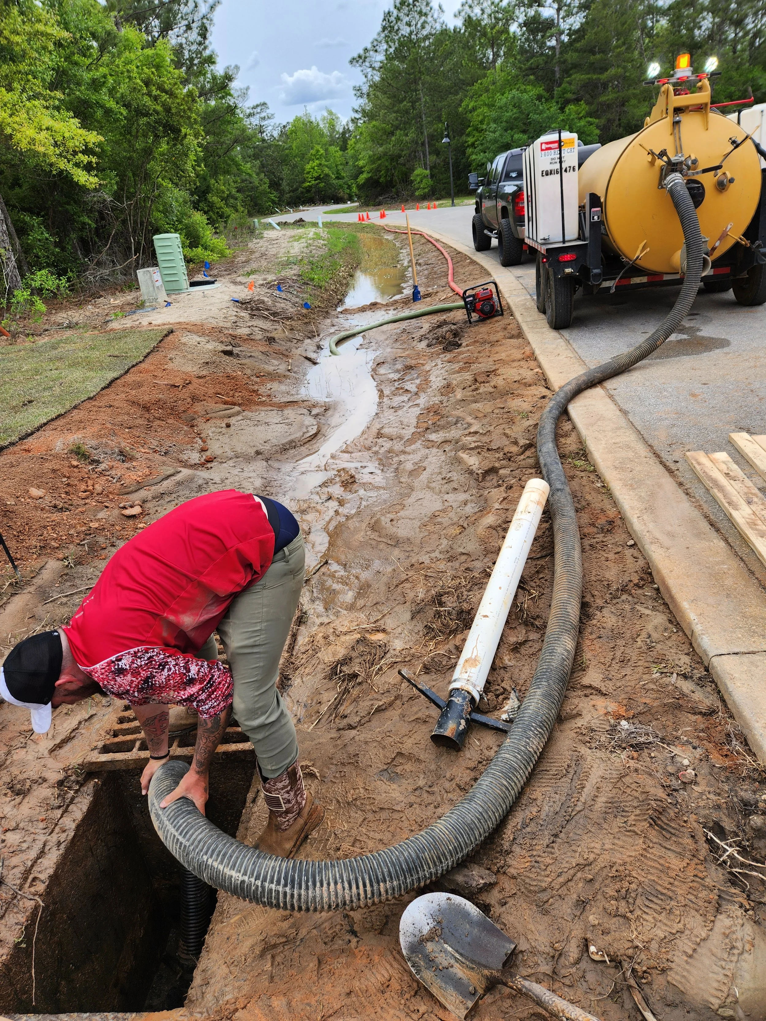 A worker installing or repairing underground drainage pipes along a street, with a large vacuum truck attached to a hose for excavation. The scene shows construction equipment, orange cones, and a natural, green background with trees.