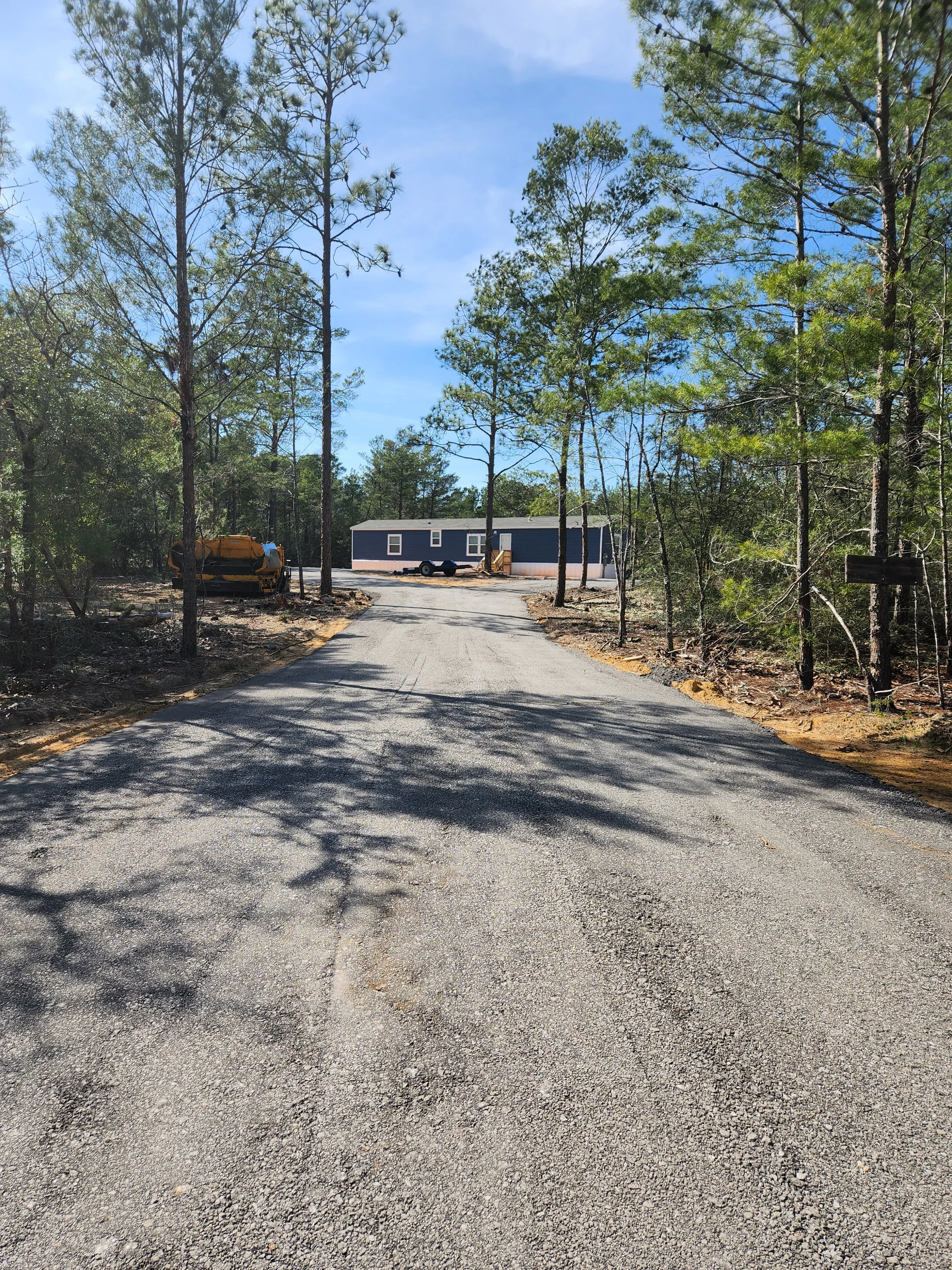 A gravel driveway leading to a small blue mobile home with white trim, surrounded by trees on a sunny day.