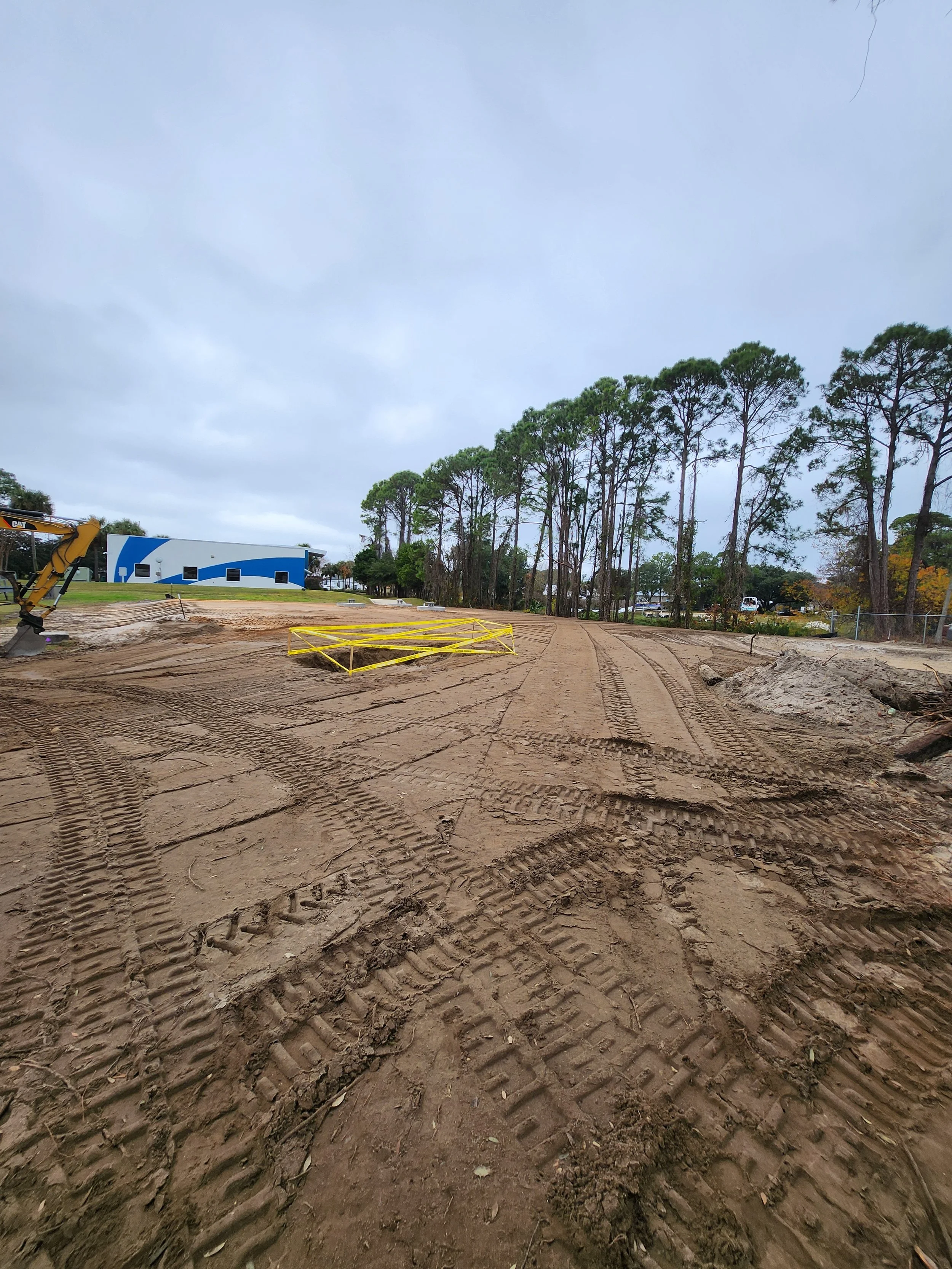 A construction site with tire tracks in the dirt, yellow safety barrier, construction equipment on the left, and trees and a building in the background under cloudy sky.