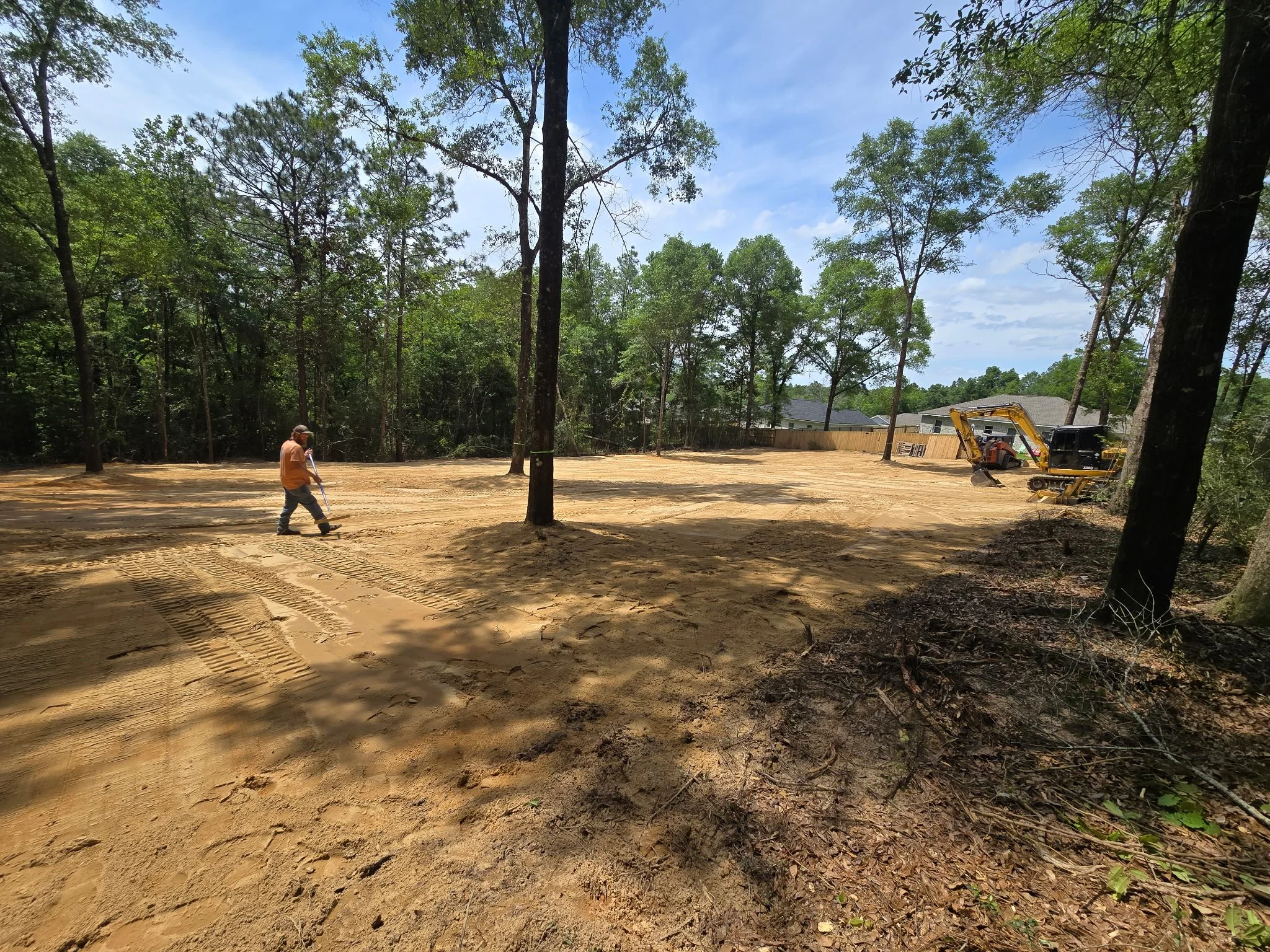 Construction site in a wooded area with a worker leveling the ground and heavy machinery parked in the background.