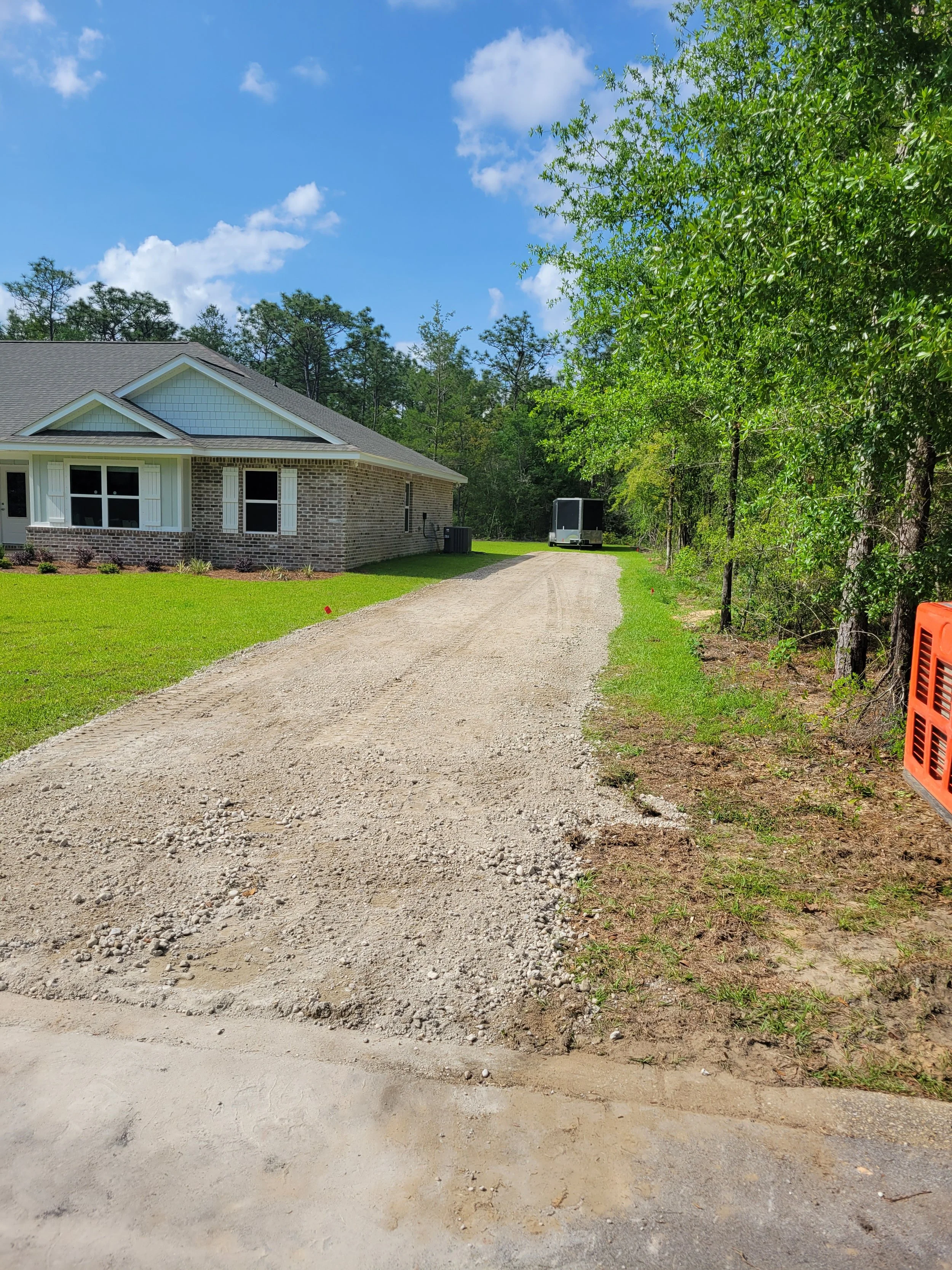 A gravel driveway in front of a house with a brick exterior and white trim, surrounded by a grassy lawn with landscaping, and trees on the right side, under a partly cloudy blue sky.