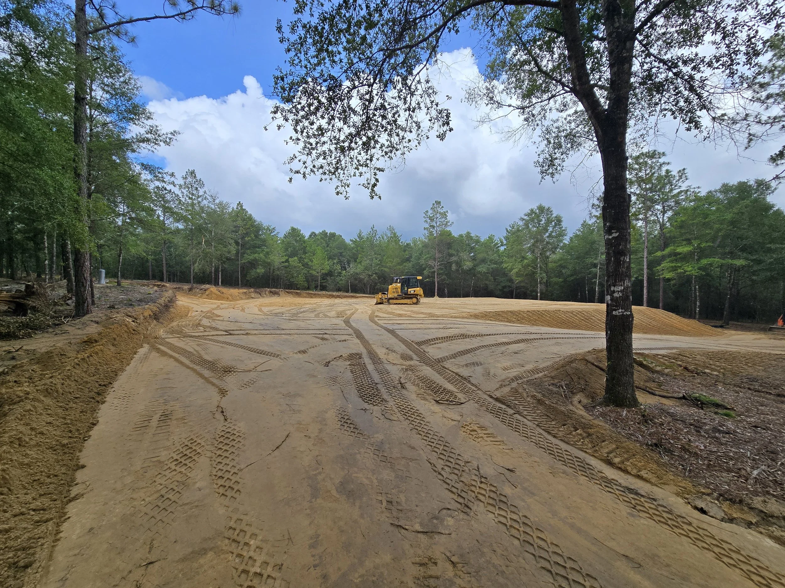 A construction site in a wooded area with a bulldozer working on leveled dirt ground, tire tracks visible in the soil, green trees surrounding the site, and a partly cloudy sky overhead.