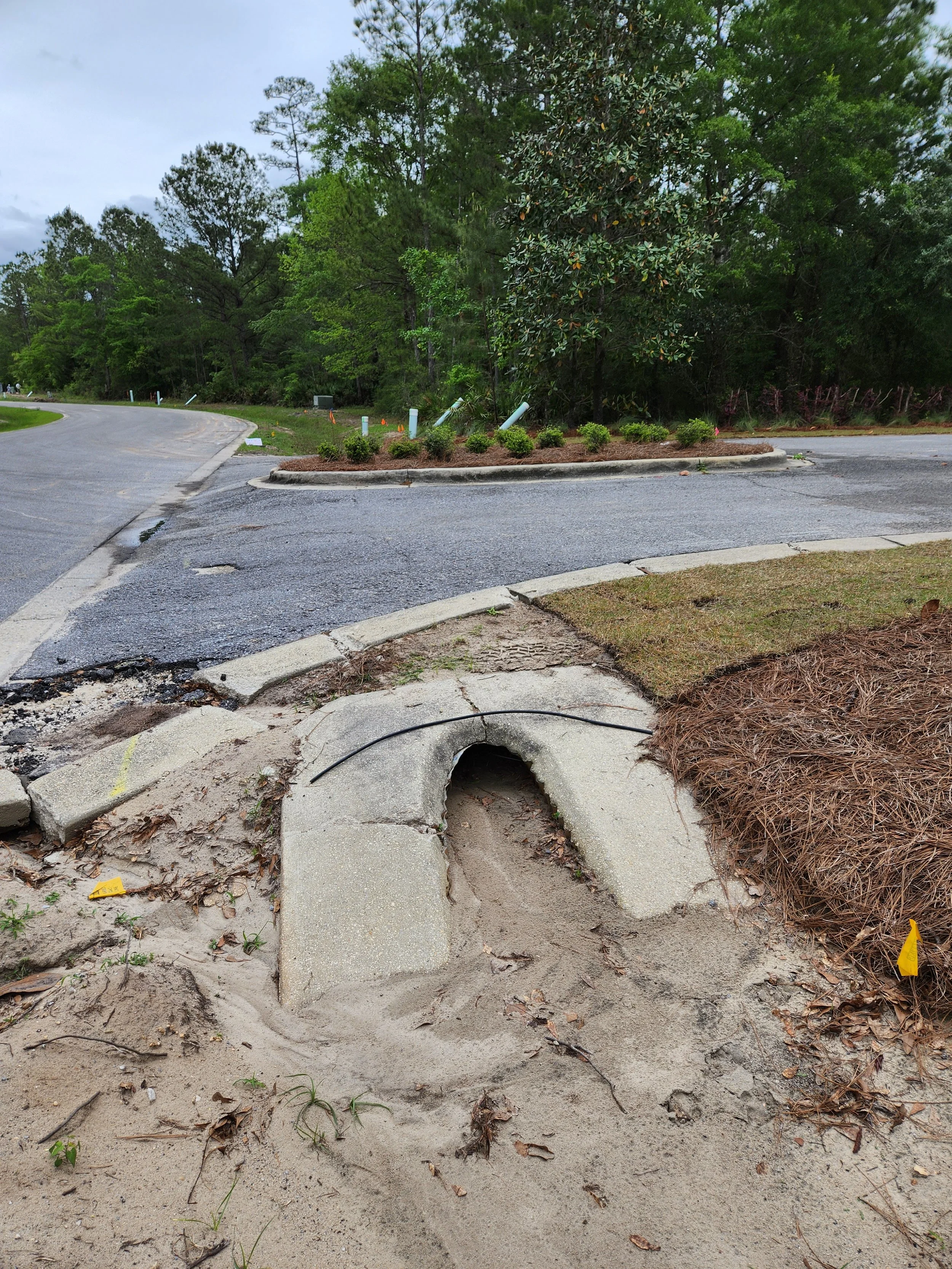 A damaged curb and sidewalk at an outdoor location, with a construction site or roadwork area in the background, surrounded by trees and greenery.