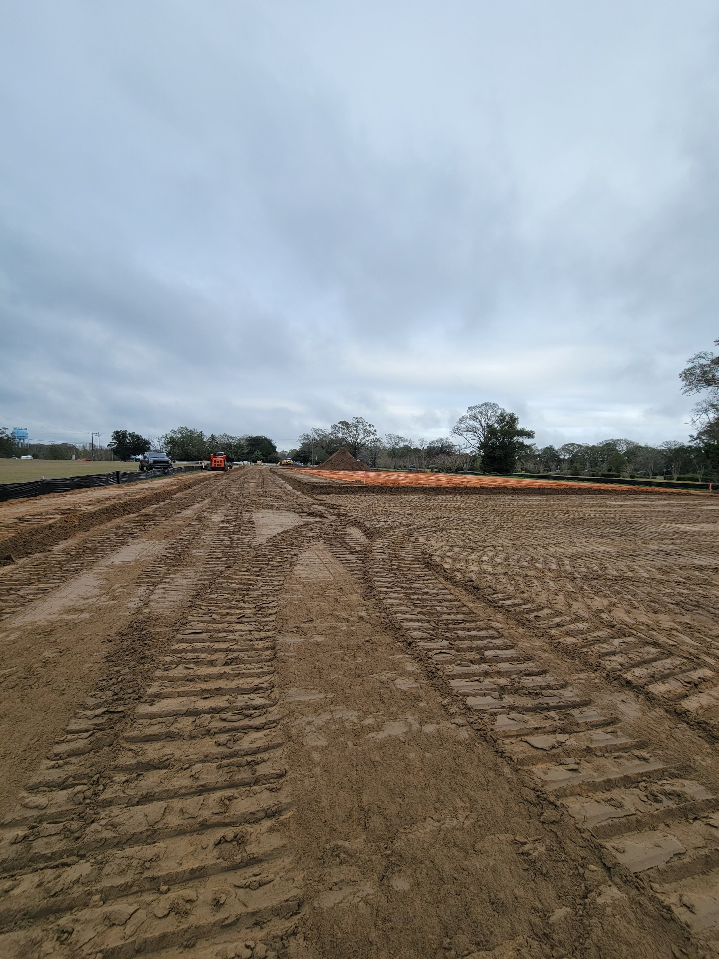 Construction site with dirt and tire tracks, construction vehicles in the distance, and overcast sky.