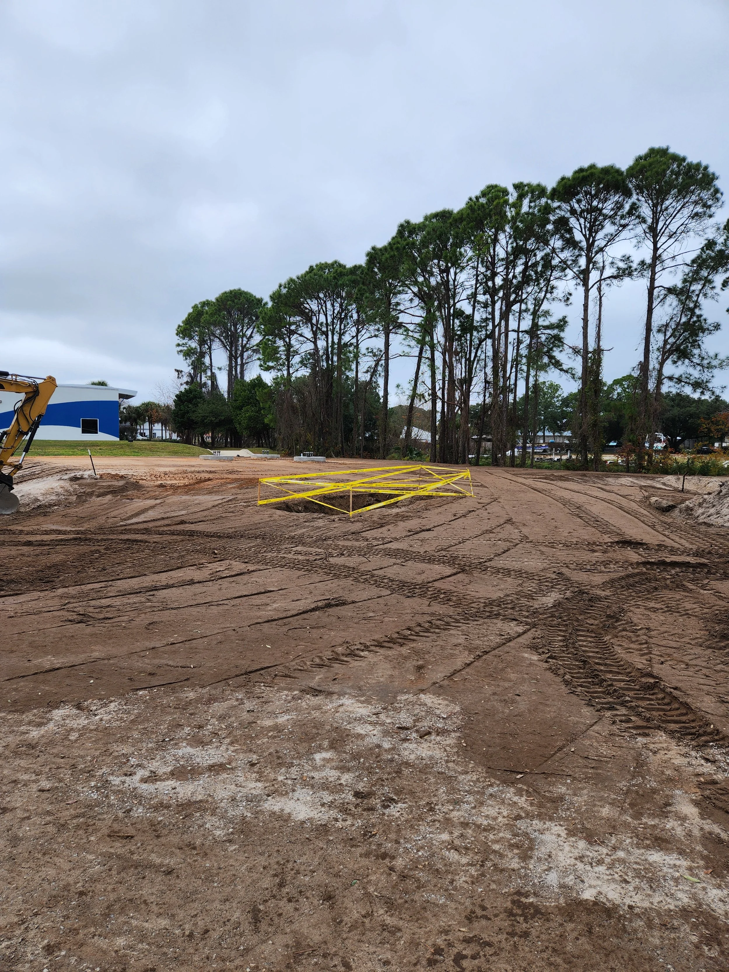 Construction site with dirt ground, yellow barricades, and a row of tall trees in the background under cloudy sky.