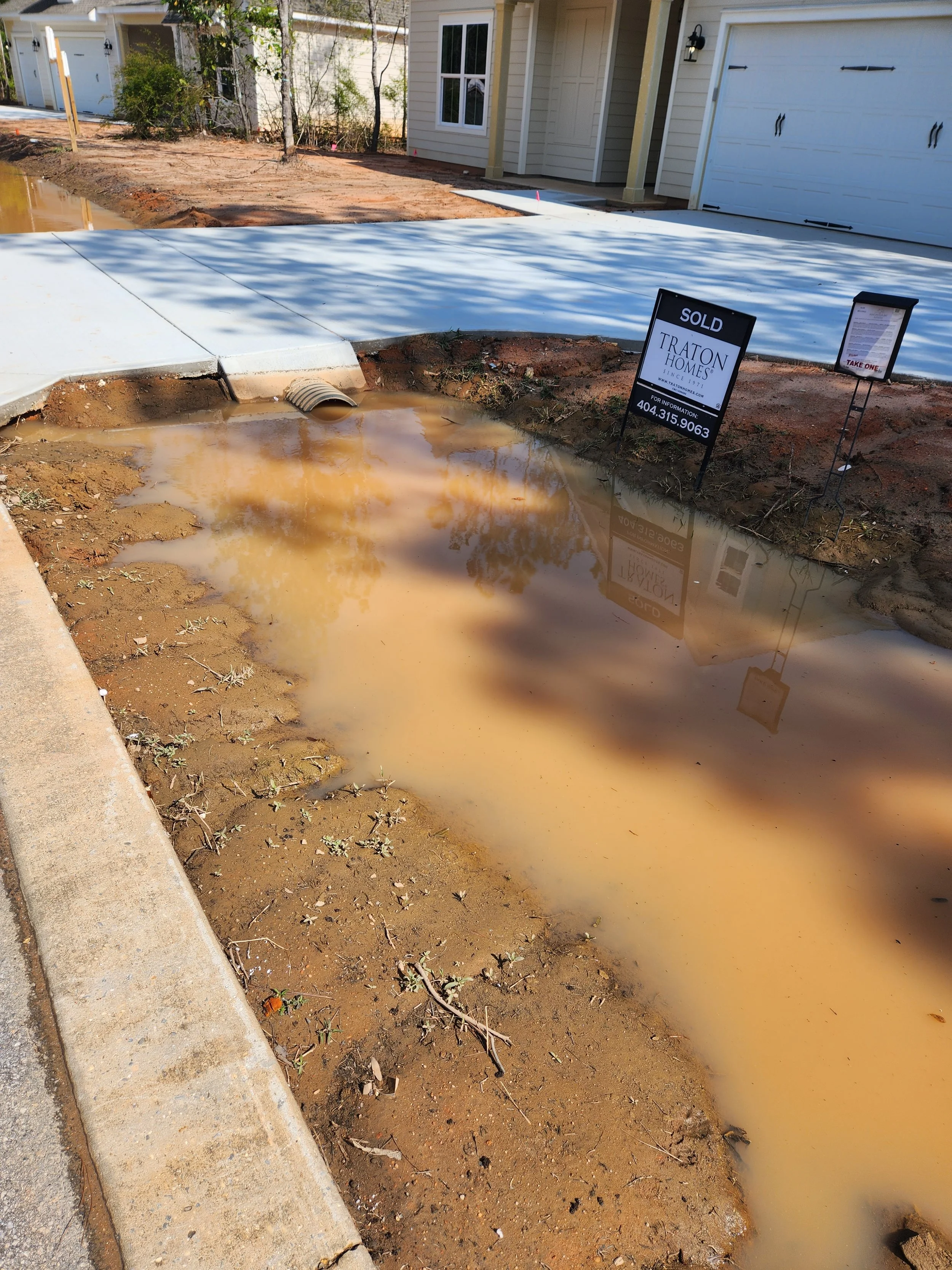 Construction site with muddy water, concrete pavement, and a house with a garage. A sign indicates the property is sold.