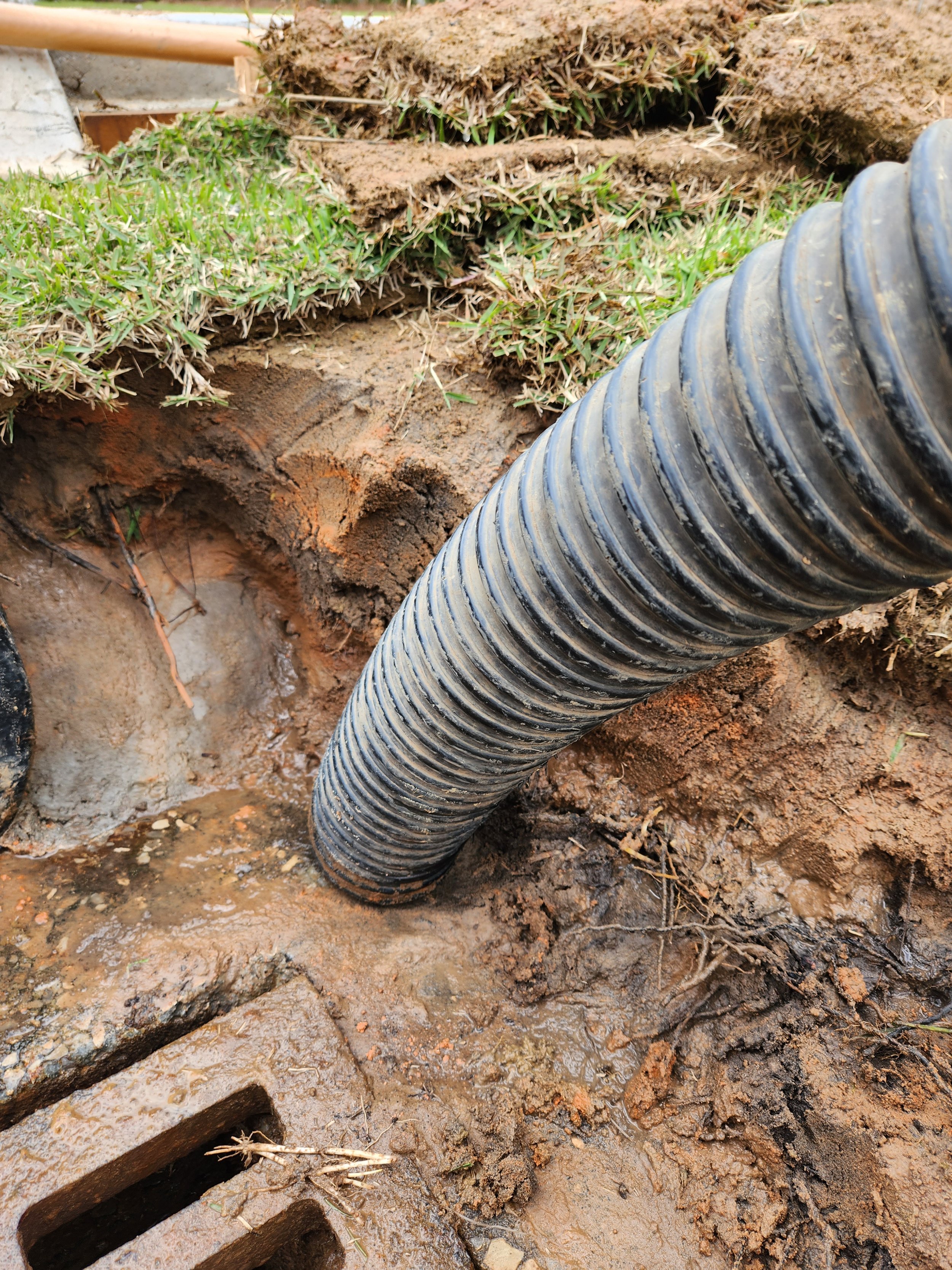 A black corrugated pipe installed underground, with earth and grass surrounding it, near a drain grate.
