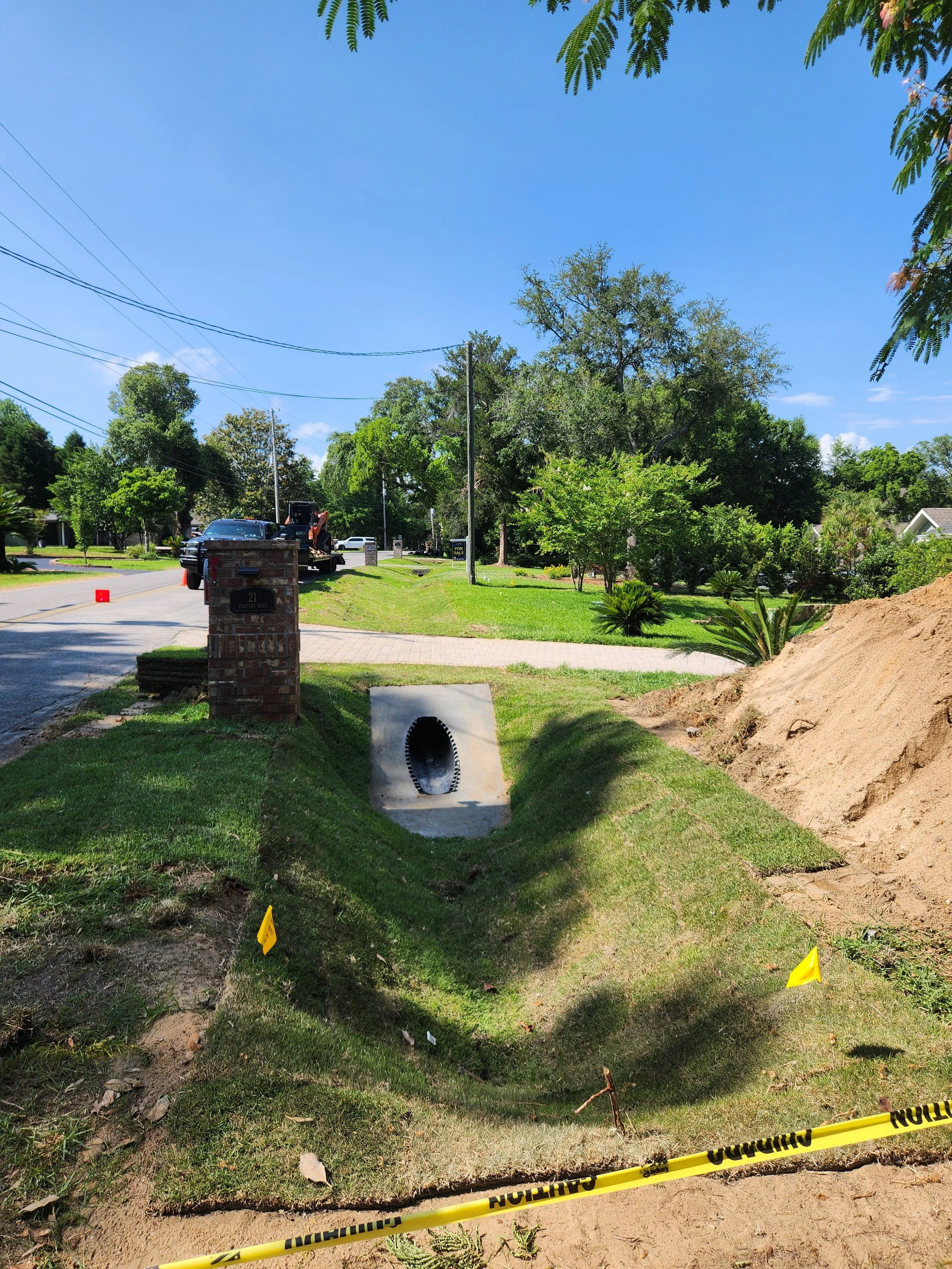 Street with ongoing sidewalk and drainage construction, yellow caution tape, orange cones, construction equipment, and green trees under a blue sky.