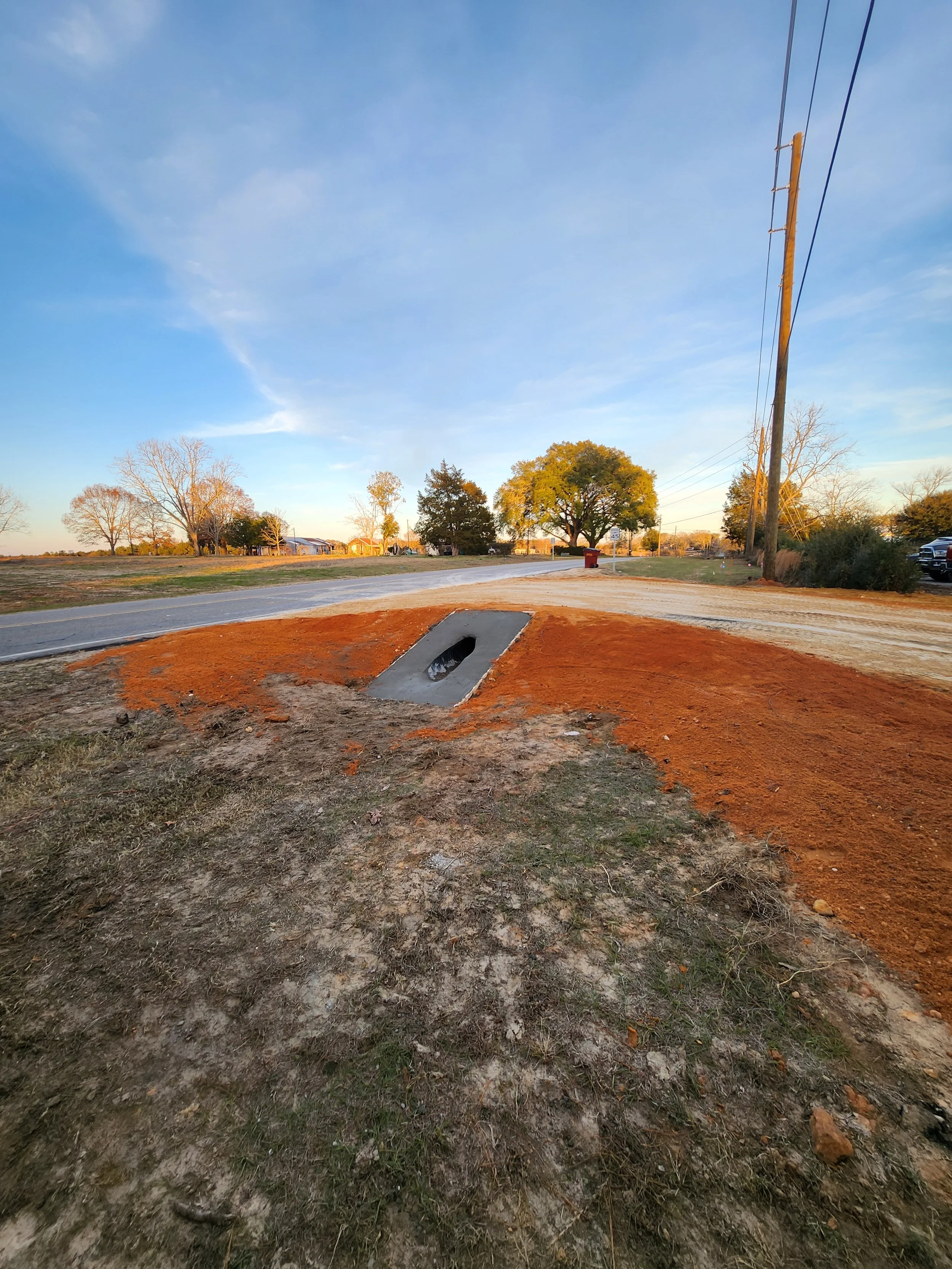 Close-up of a roadside ditch with concrete drainage structure, surrounded by soil and grass, under a clear blue sky with trees and utility poles in the background.