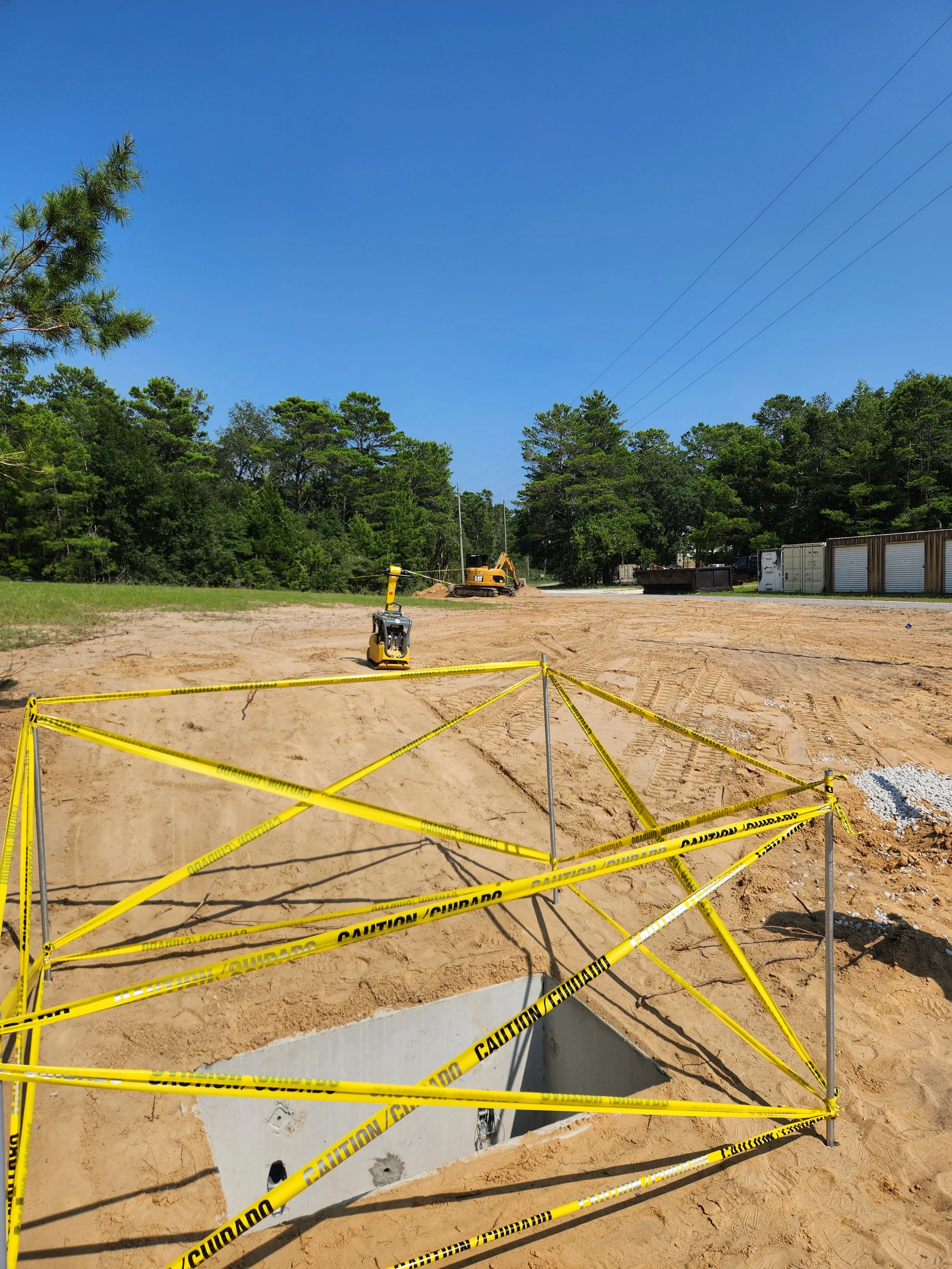 A construction site with a large open hole surrounded by yellow caution tape, construction equipment in the background, and a bright blue sky.