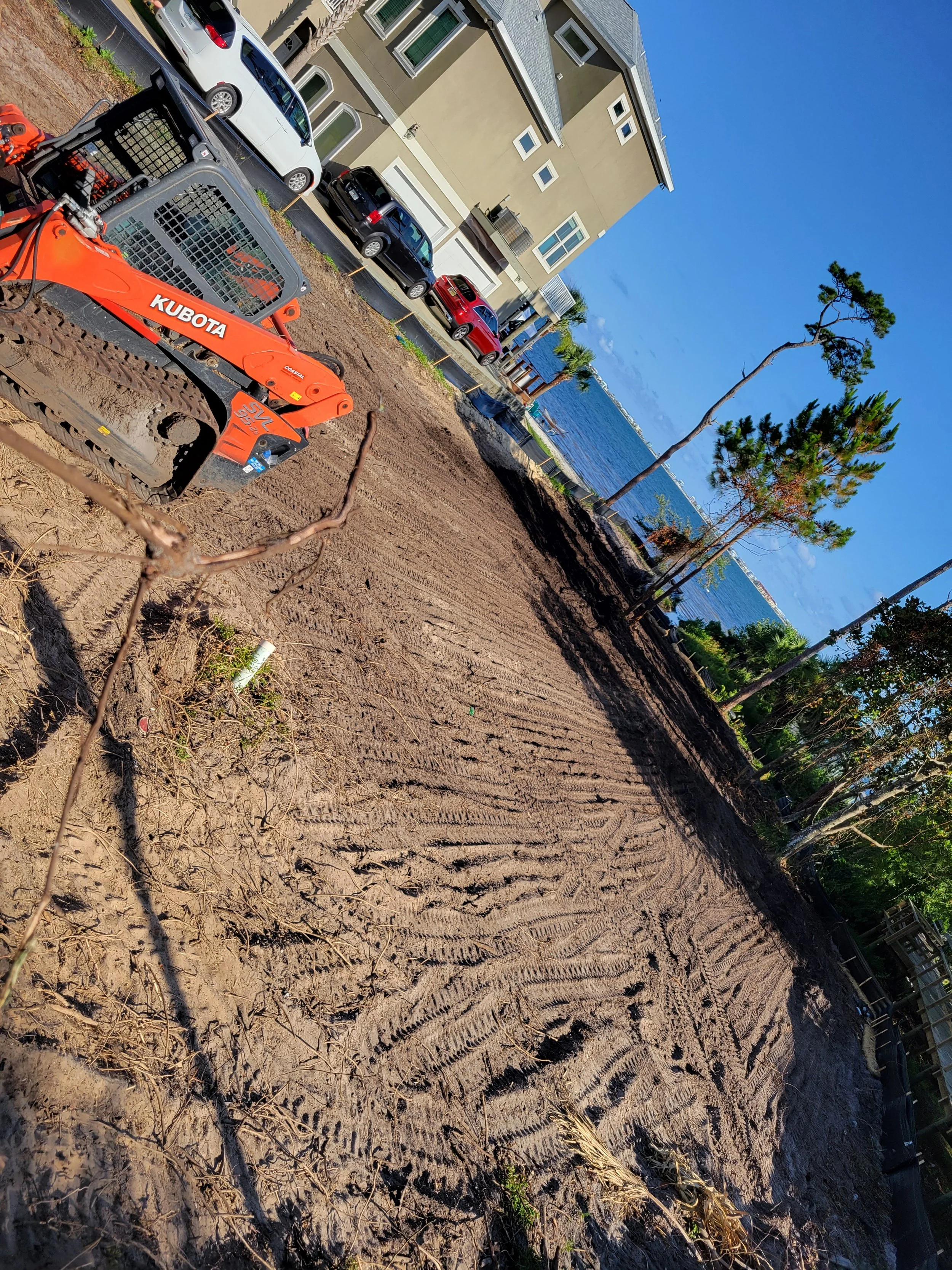 A construction site with freshly tilled dirt, tire tracks, and a Kubota mini excavator parked nearby. In the background, there are several parked cars in front of a multi-story house overlooking a body of water under a clear blue sky.