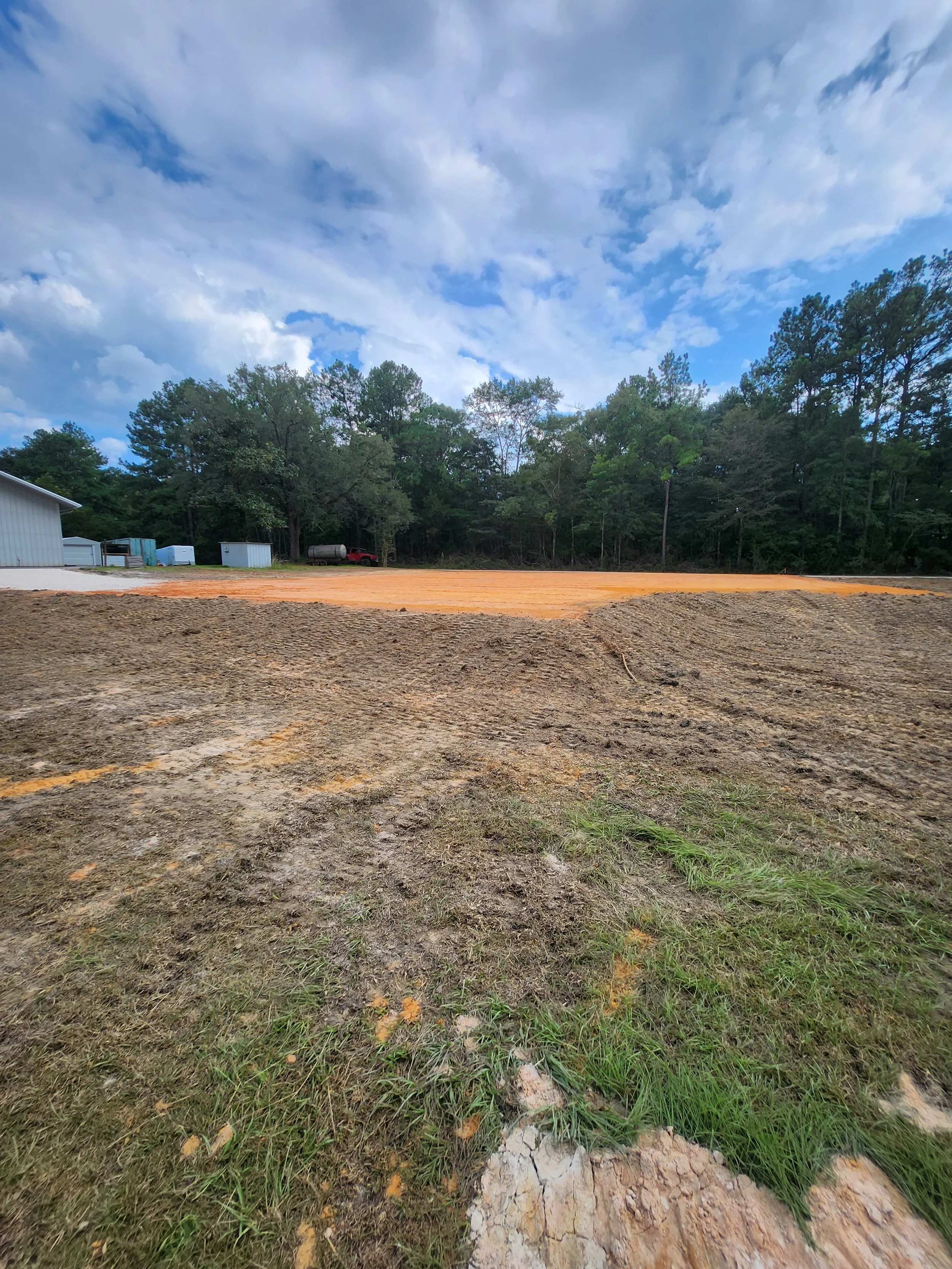 A construction site with cleared land, some grass and dirt in the foreground, and a line of trees in the background under a partly cloudy sky.