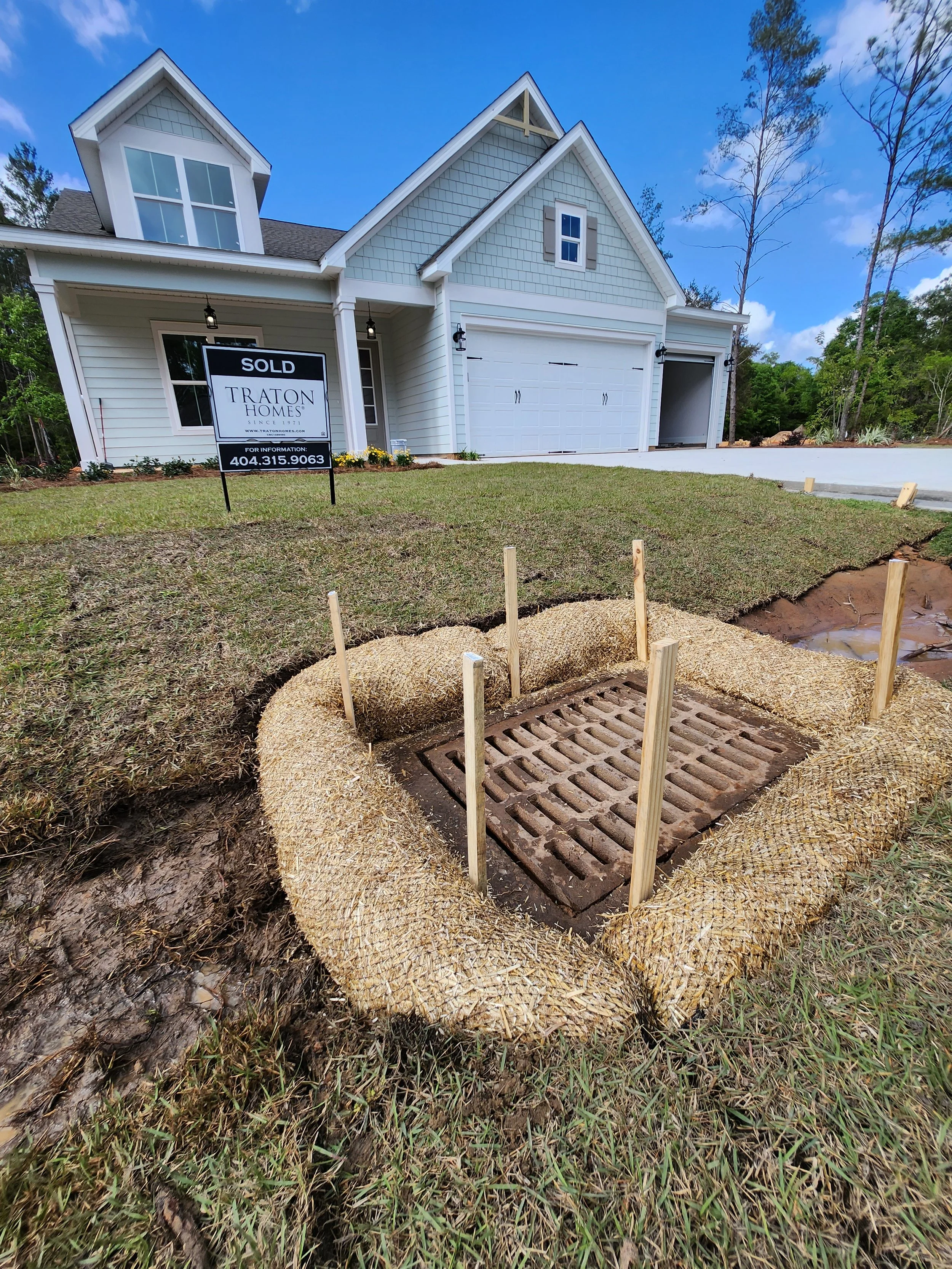 Newly landscaped front yard of a house with a storm drain surrounded by straw, wooden stakes, and a lawn with a sold sign in front of the house.