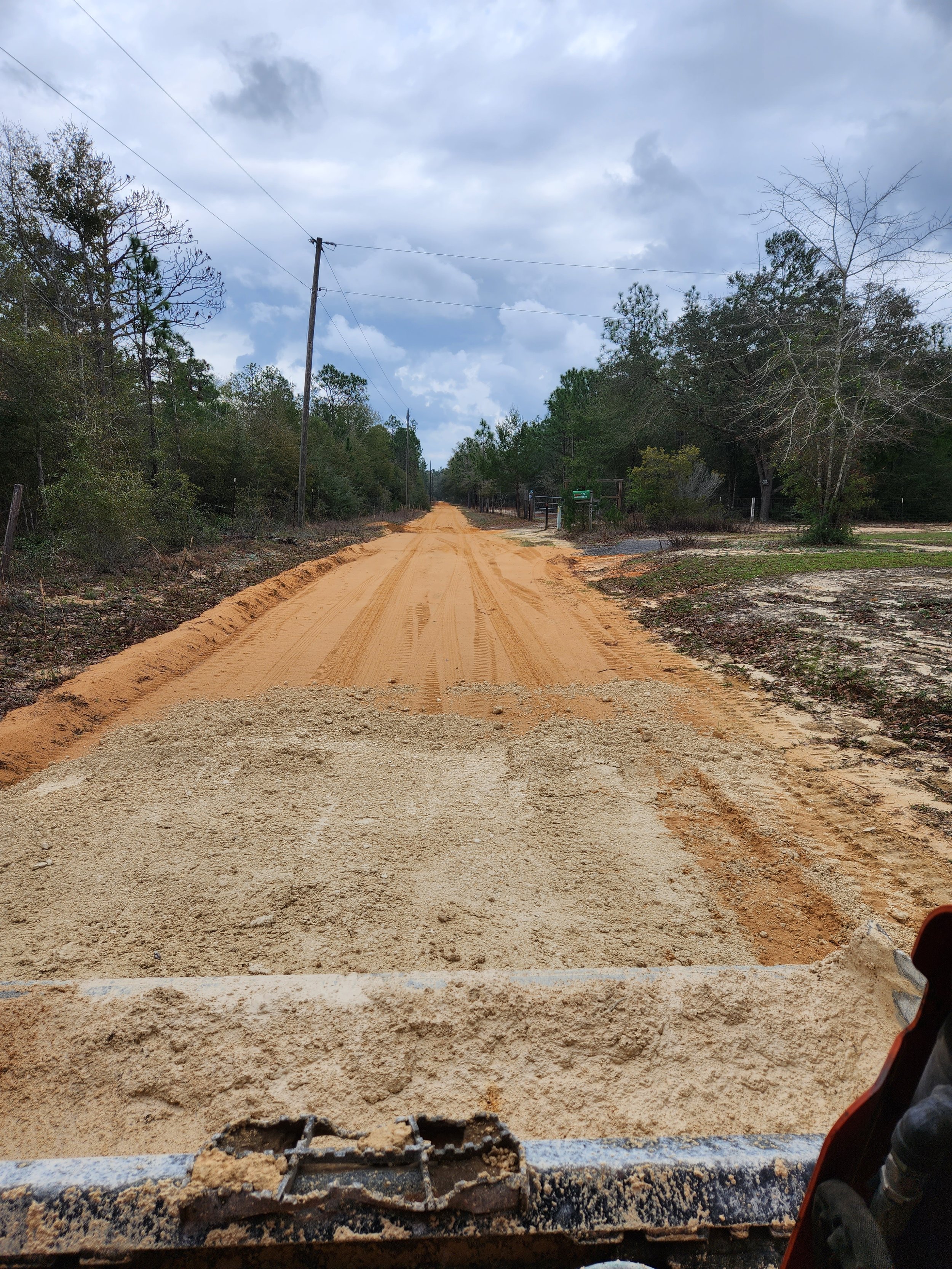 A dirt road under cloudy skies, with power lines, trees, and some structures on the sides, taken from the front of a vehicle.