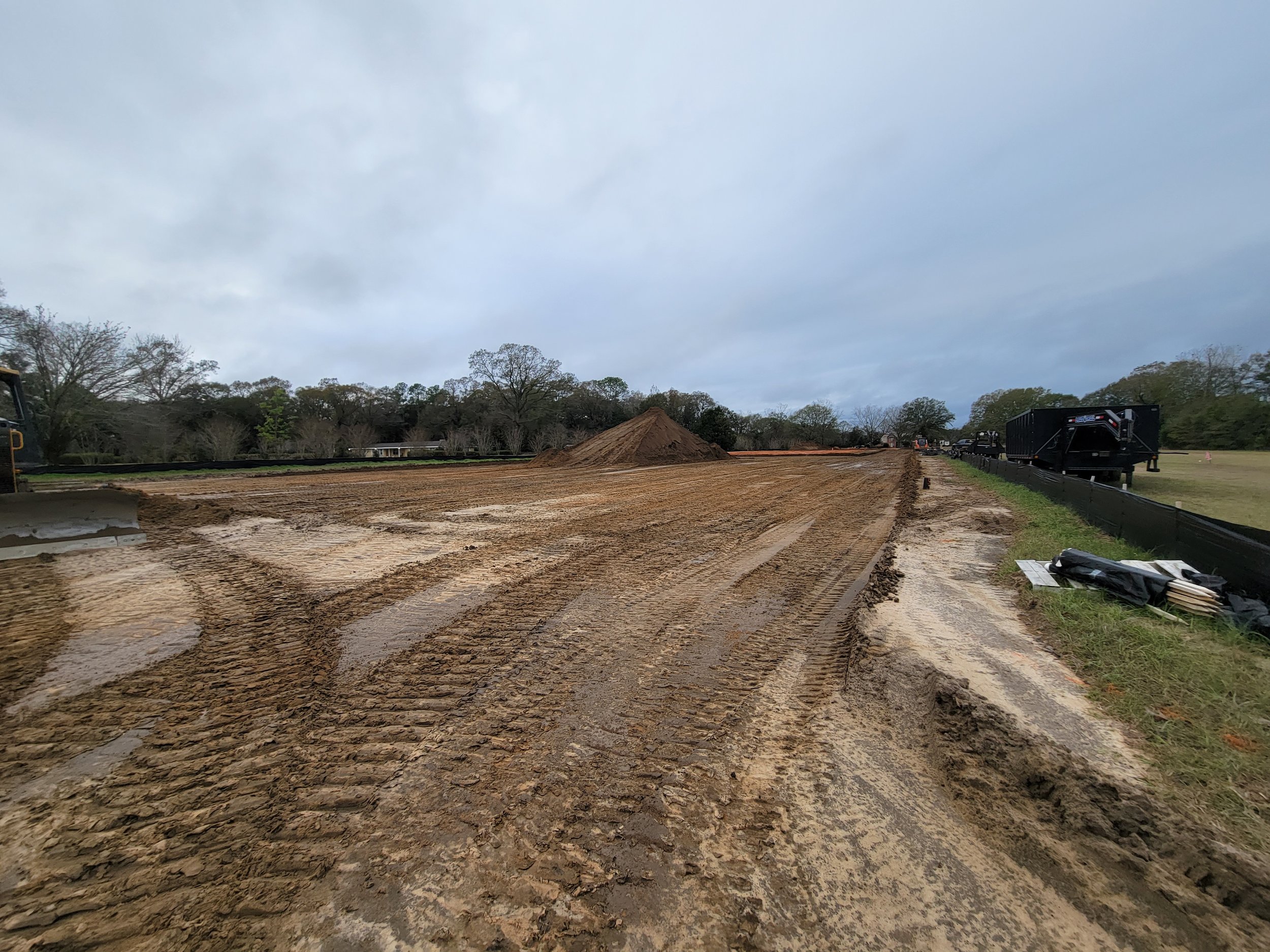 A construction site with muddy ground, construction vehicles, and a dirt mound in the background, with cloudy skies overhead.