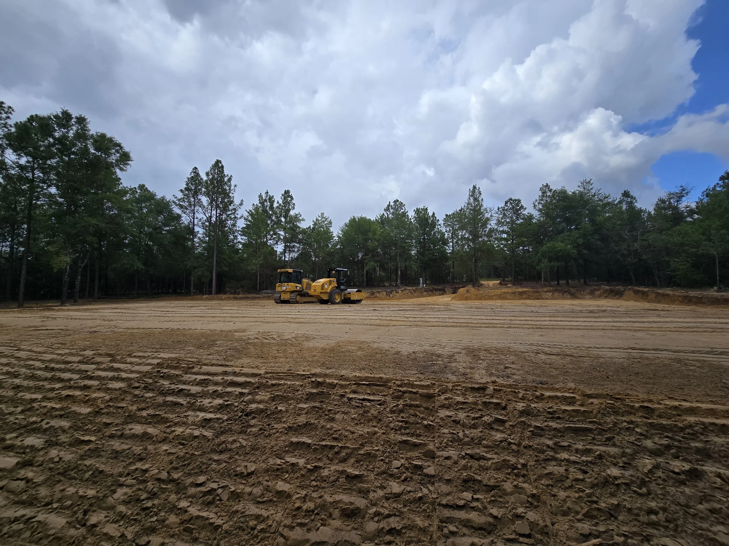 Construction site with two yellow rollers on cleared dirt ground surrounded by trees and cloudy sky.