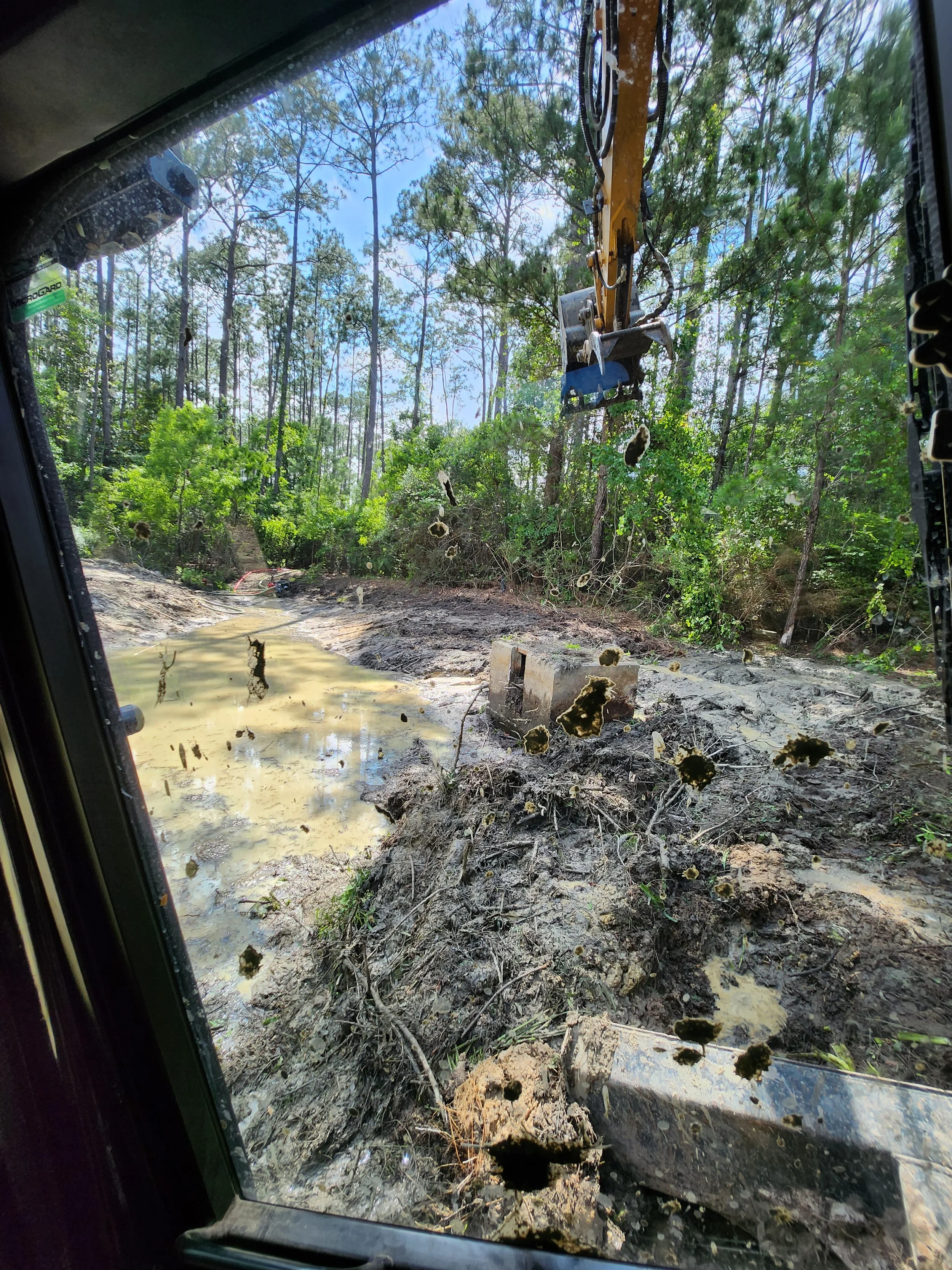 View from inside a construction vehicle cabin of a muddy, uneven dirt road with tire tracks, a small pool of water, muddy debris, and surrounded by trees and greenery. clearing ponds 