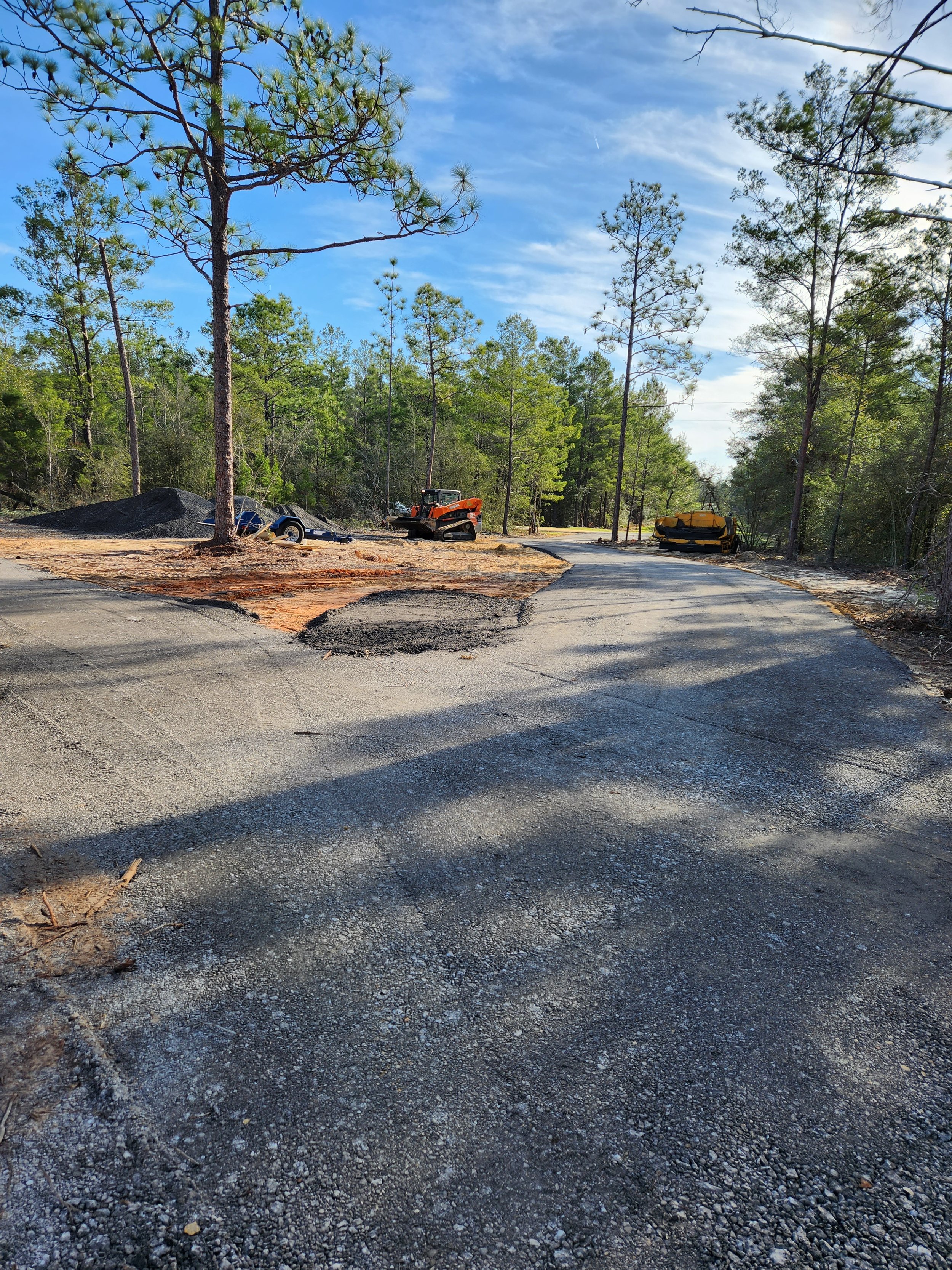 Road construction site in a forested area with paving equipment, trees, and piles of gravel