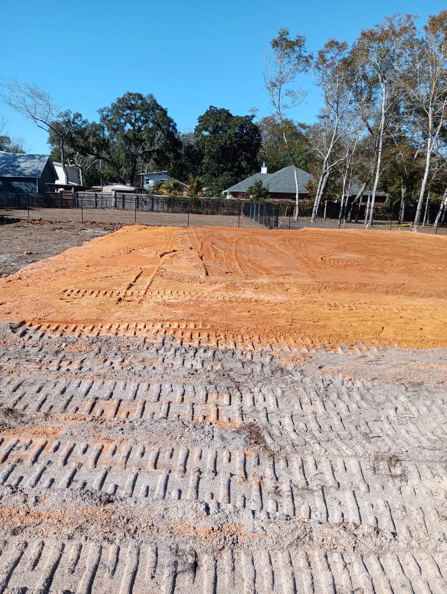A construction site with freshly laid orange-colored dirt and tire tracks, surrounded by a fence and residential houses in the background under a clear blue sky.