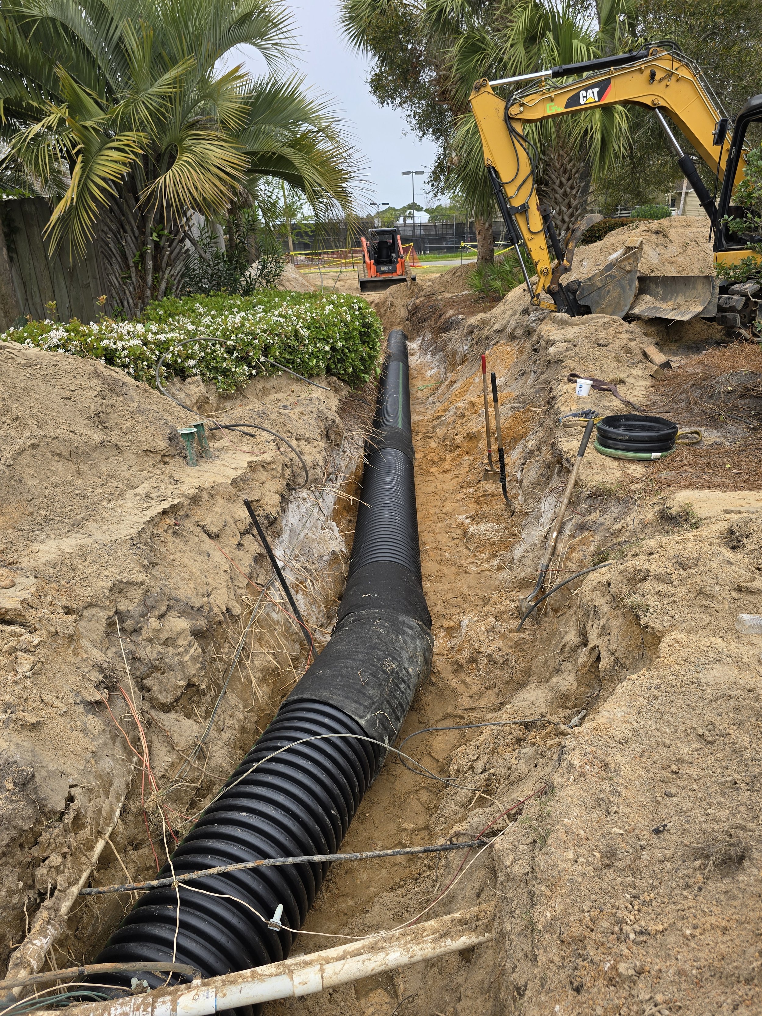 Construction site with a large black underground pipe being installed in a deep trench, with an excavator nearby, surrounded by palm trees and bushes.