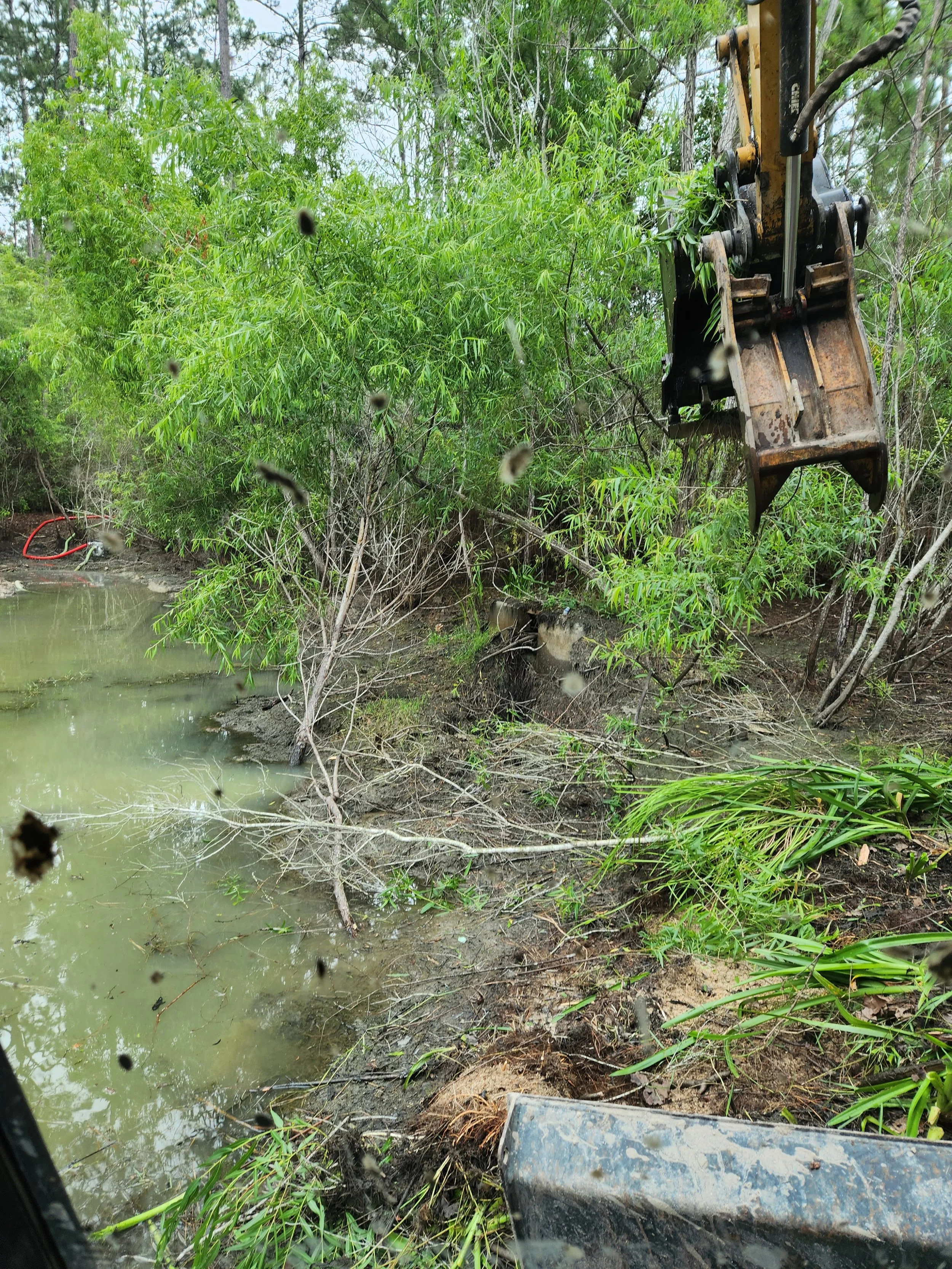 View from a vehicle window showing a muddy shoreline with green plants and bushes, a body of water, construction equipment with a yellow hydraulic arm, and some fallen branches and debris. clearing ponds 