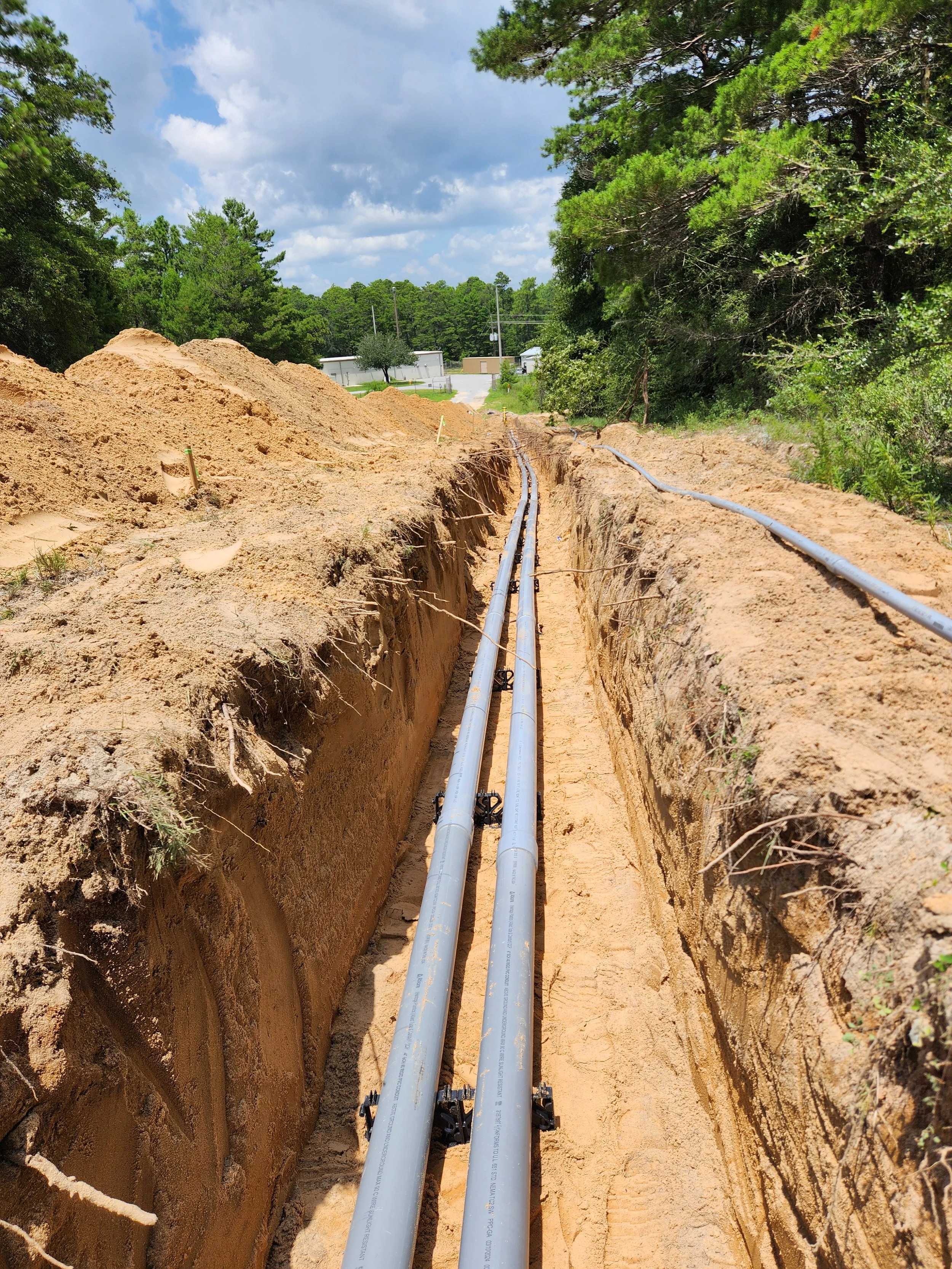 Construction site with two parallel utility pipes laid in a trench in a rural area with trees, cloudy sky, and some buildings in the background.