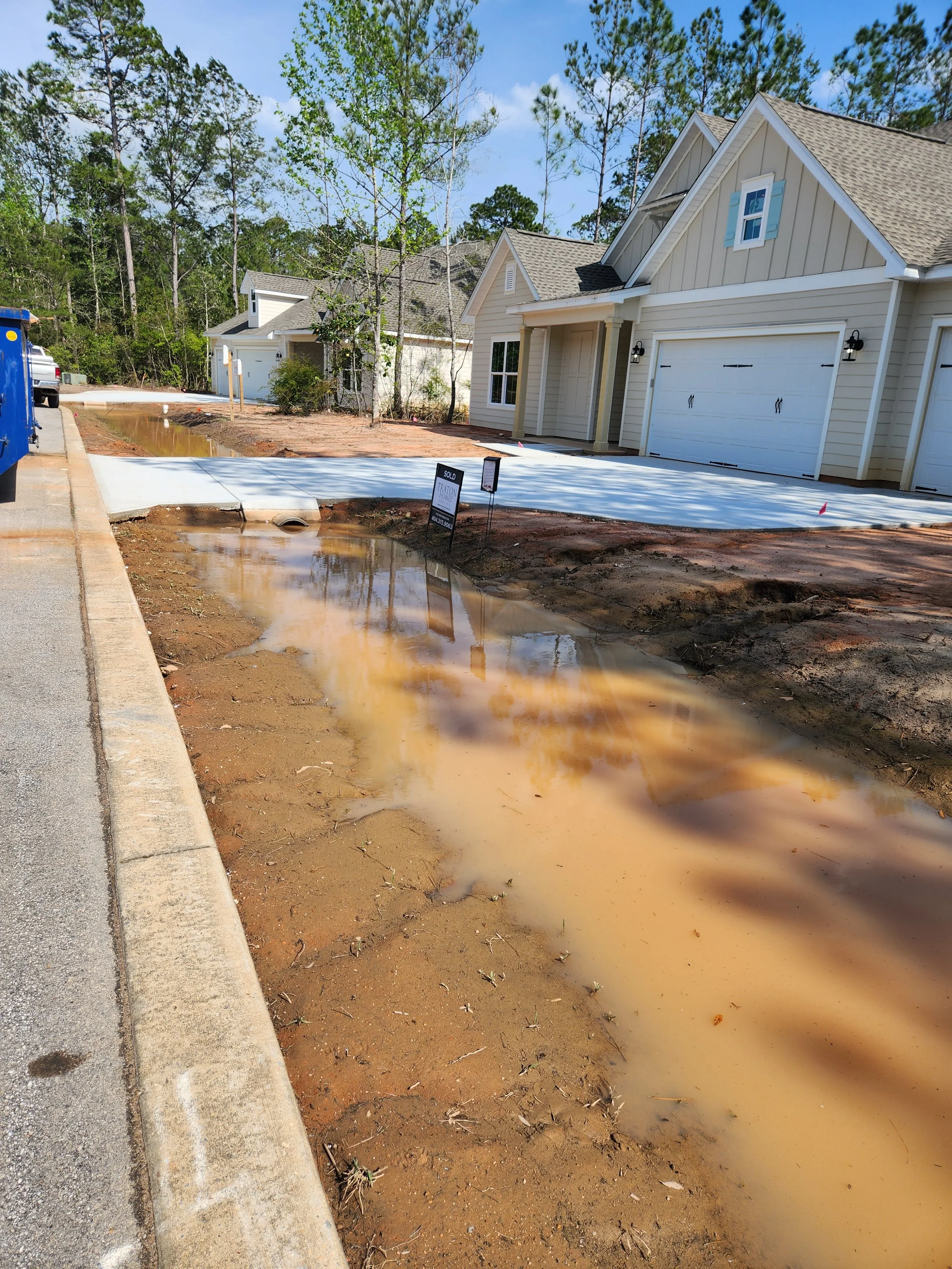 A newly constructed suburban house with a garage, construction signs, a concrete driveway, and a muddy puddle of water in the front yard, with trees and a blue sky in the background.