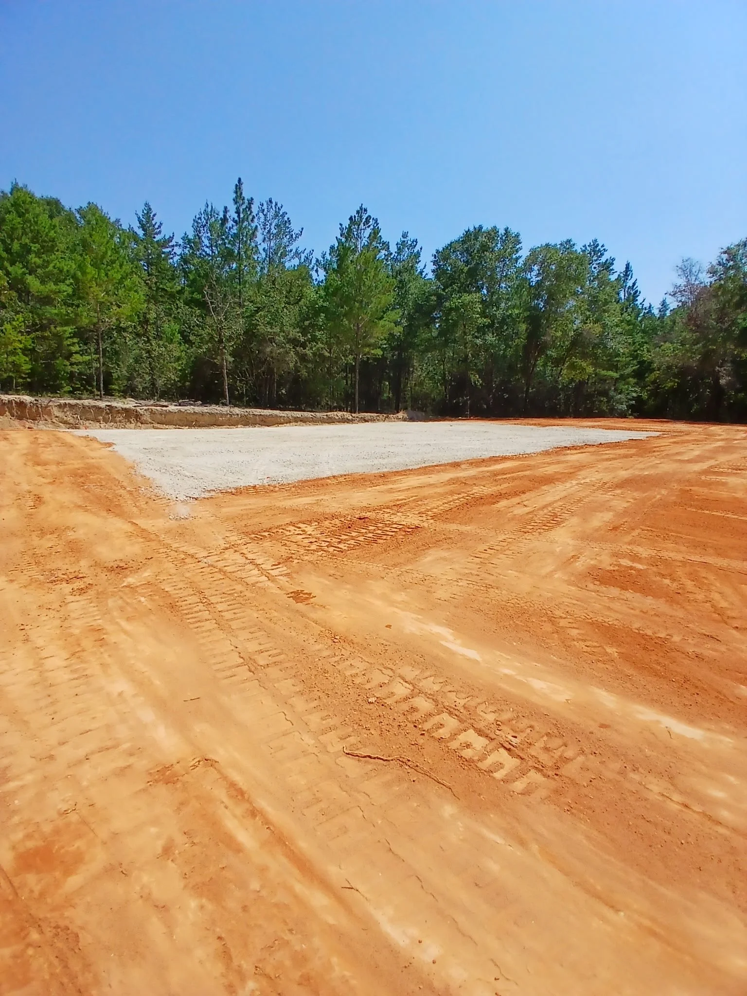 Construction site with cleared land, orange dirt, and trees in the background under a clear blue sky.