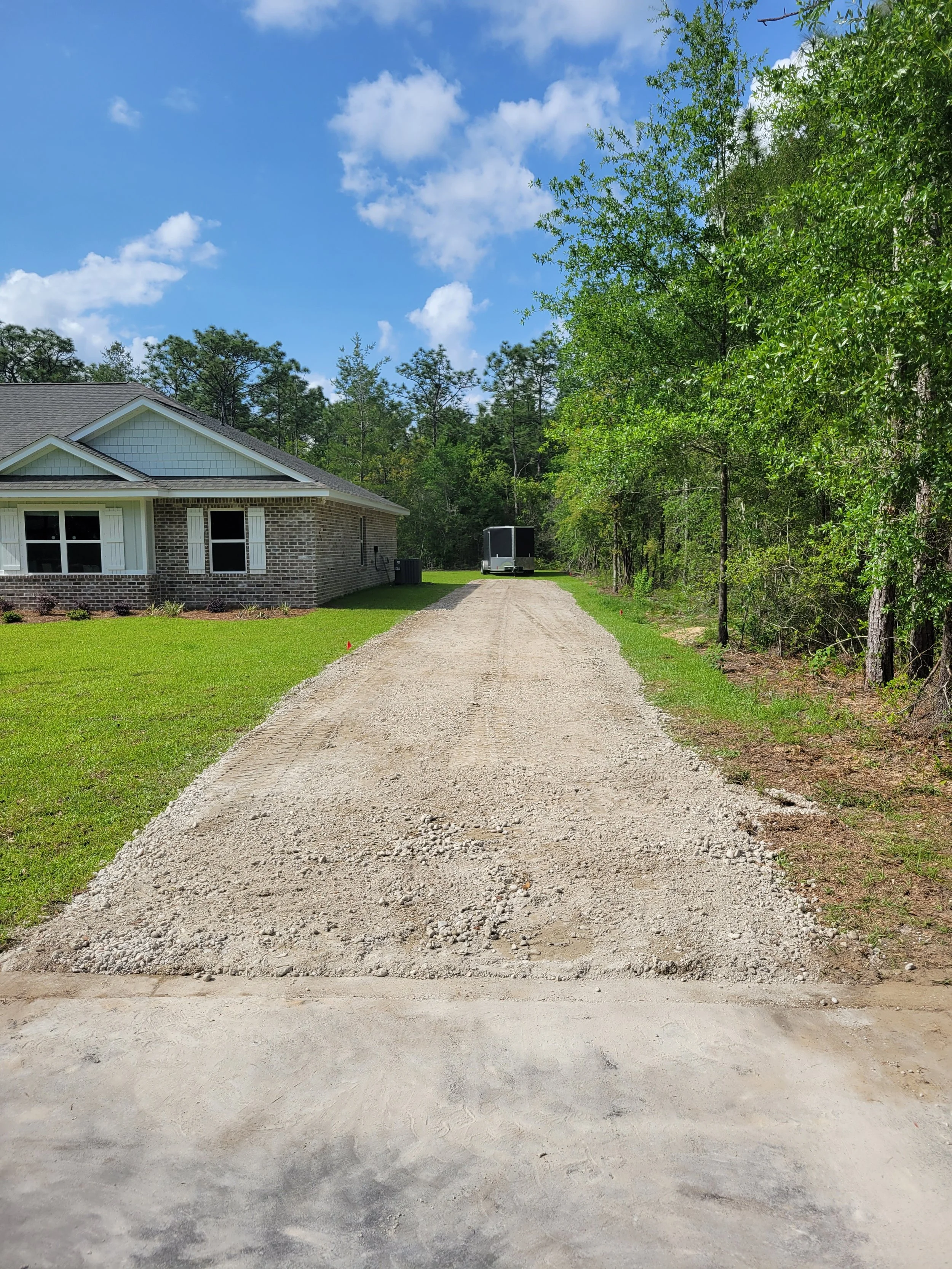 A gravel driveway leading to a house with a brick exterior and white window shutters, surrounded by green grass and trees under a partly cloudy sky.
