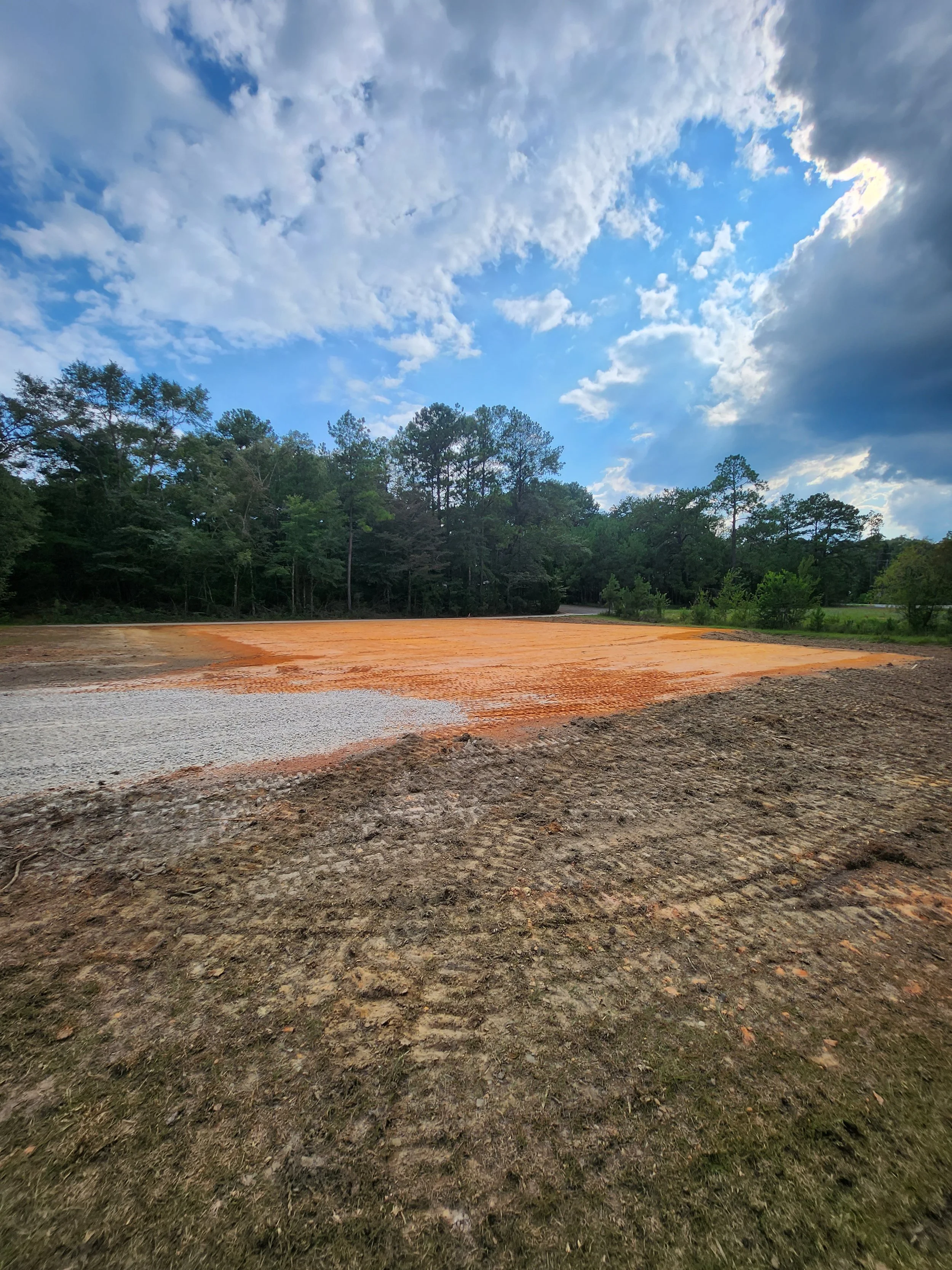 A construction site with a leveled area of orange and white materials, surrounded by trees under a partly cloudy sky.